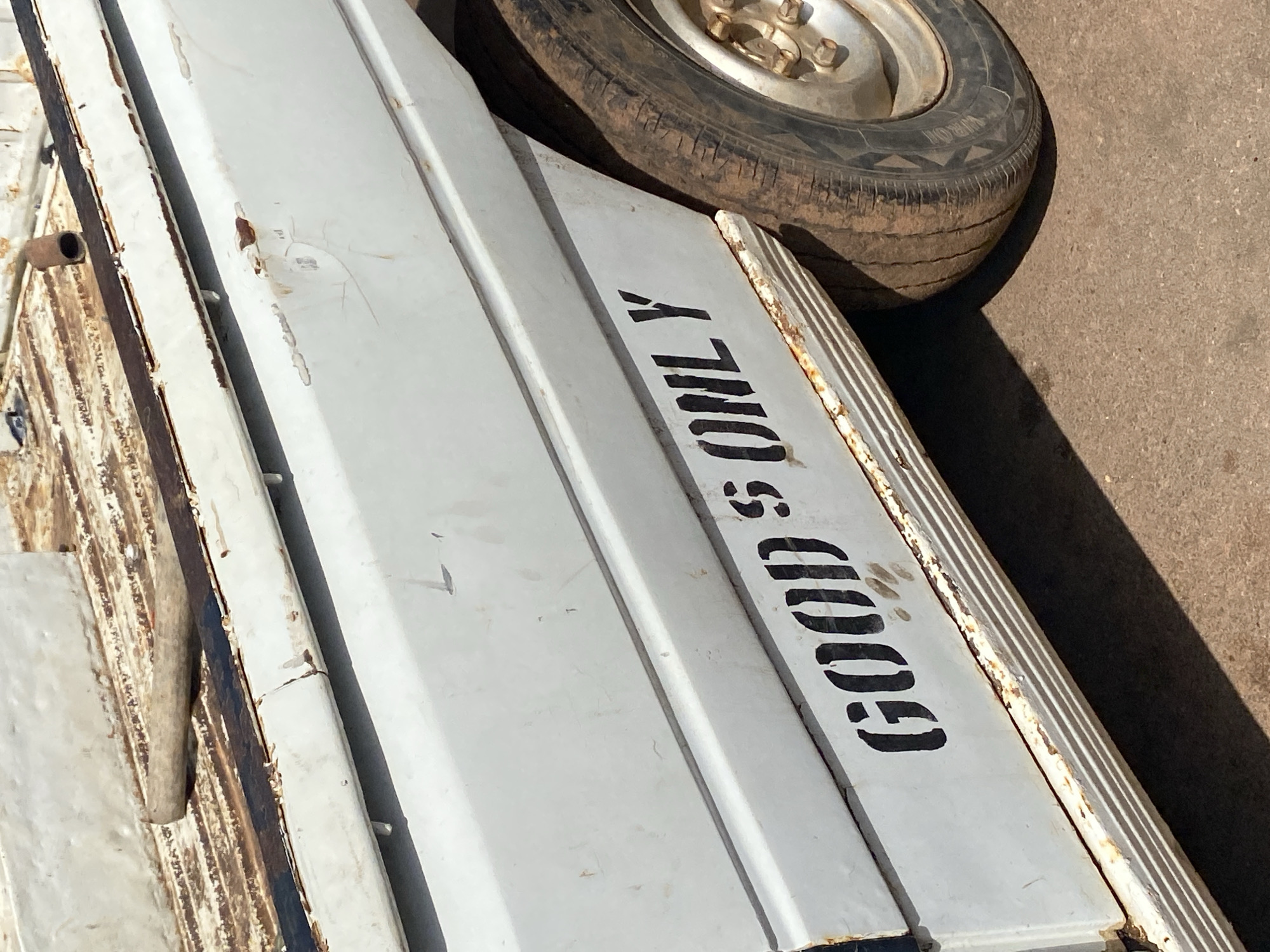 The image shows the side of a white, rusty pickup truck parked outdoors on a dirt or asphalt surface. Stenciled in black capital letters on the truck's side are the words "GOODS ONLY". The truck has a tire visible on the right side of the frame. The scene appears to be in Gura Top, Nigeria, indicated by the context provided. The lighting suggests it is daytime. No people are visible in the image.