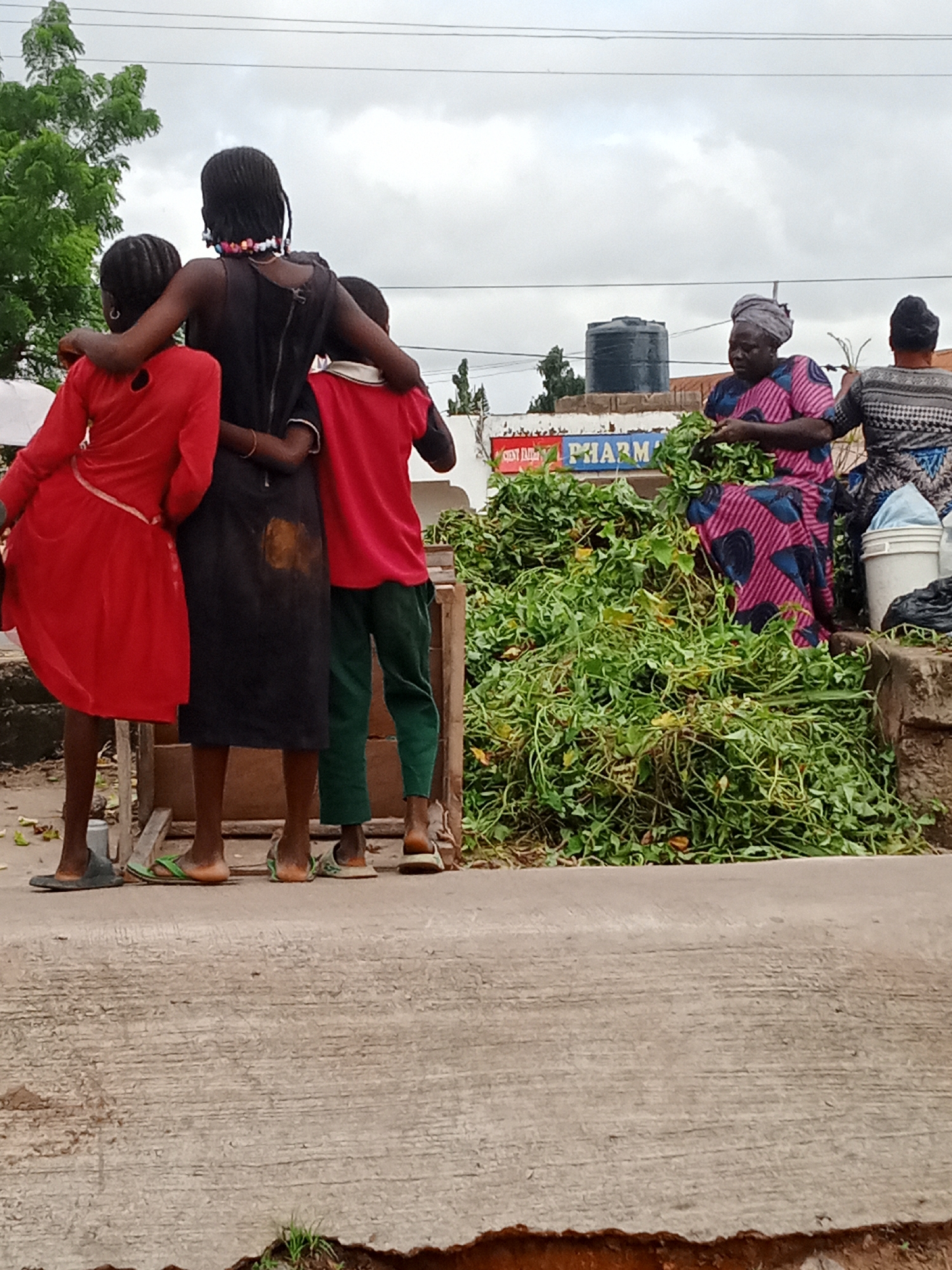 In an outdoor urban setting, three children stand shoulder-to-shoulder with their arms around each other. The child in the middle wears a black dress, while the child on the left is dressed in red, and the child on the right wears a red shirt and green pants. Behind them, two women are visible. One woman, wearing a patterned pink and blue dress and a headscarf, is handing greens to another woman who is partially visible and appears to be holding some greens as well. A large pile of leafy green vegetables is situated in front of the women. In the background, a dark blue water tank is mounted on a structure. A sign with red and blue lettering, reading "PHARM," is also visible on a building. The ground appears to be a concrete surface, with some debris and vegetation present. The sky is overcast.