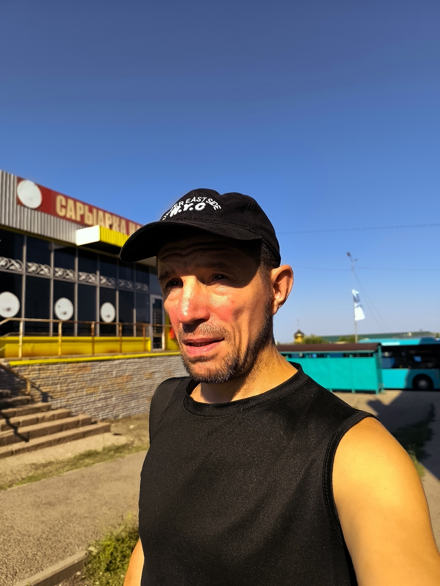 In Qarağandı, Kazakhstan, a man wearing a black sleeveless shirt and a black baseball cap stands outdoors. The cap features white text that reads "UPPER EAST SIDE N.Y.C.". He has a light beard and his face is illuminated by sunlight, casting shadows. Behind him, a building with a red and white striped facade and a prominent yellow trim is visible. The building has several large windows and an entrance. To the right of the building, a turquoise bus is parked. The ground in front of the building is paved, and there are stone steps leading up to an elevated walkway. The sky above is a clear, bright blue.