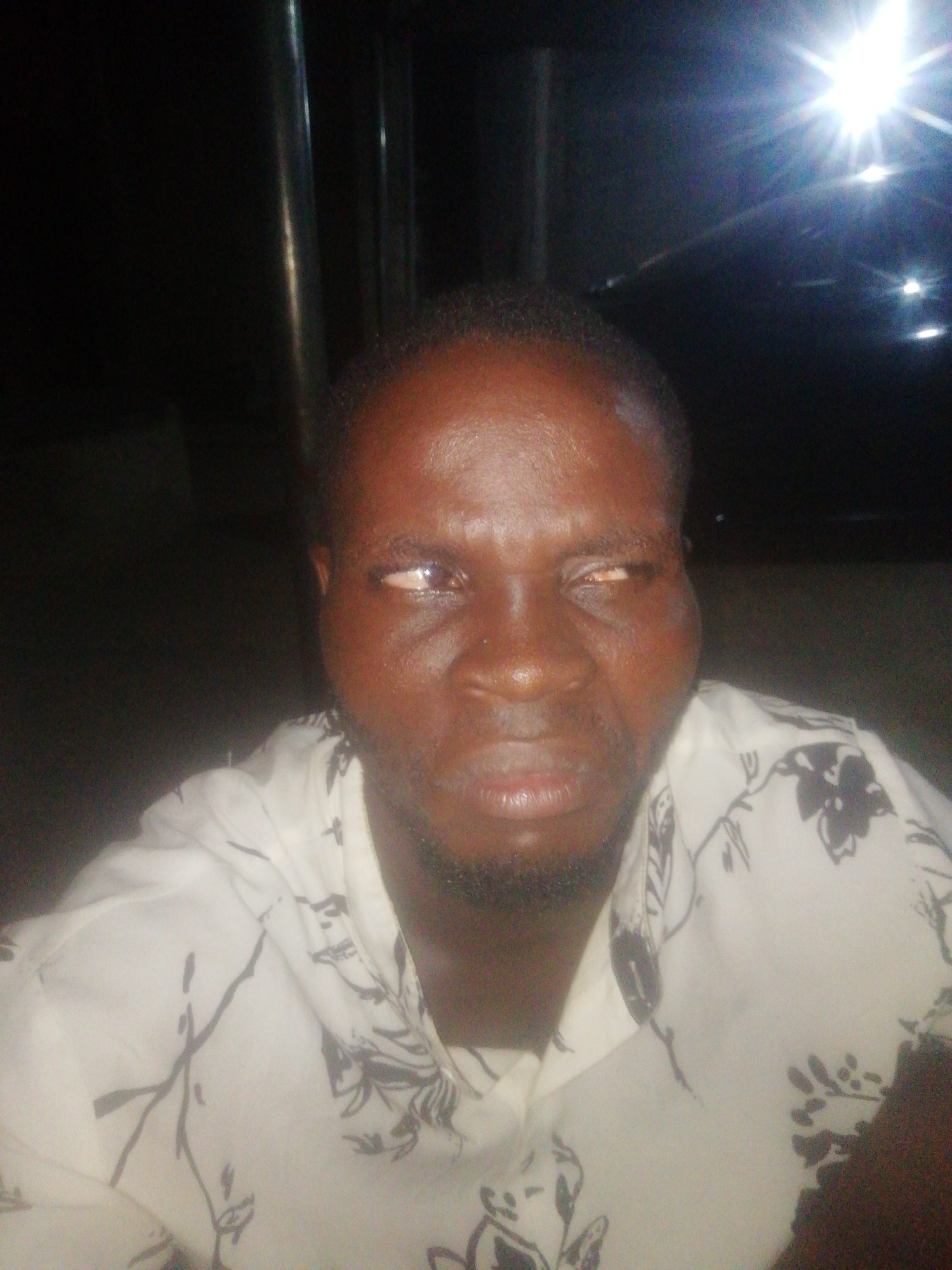 A dark-skinned man with close-cropped hair is centered in the frame. He is wearing a light-colored shirt with a dark floral pattern. His eyes are noticeably misaligned. The location is not evident.