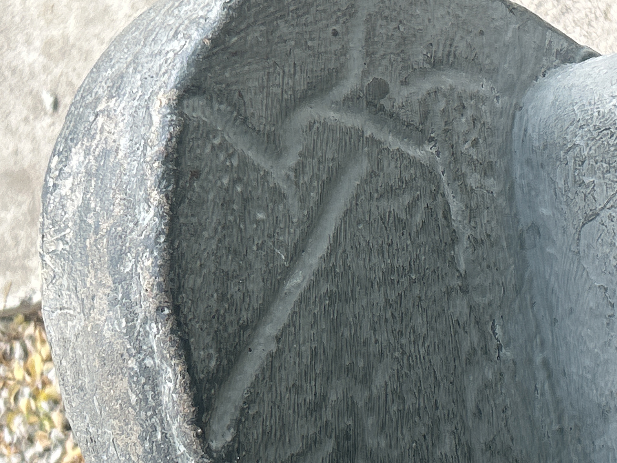 The image shows a close-up of a grey, textured surface with visible striations. There are lighter-colored, raised lines that form a pattern, appearing to be carved or embedded into the surface. The surface is curved and appears to be part of a larger object, possibly a statue or architectural element. In the upper left corner, out-of-focus foliage with autumnal colors is visible. The background is a light-colored, textured surface, likely concrete or pavement. The image does not contain any discernible people, actions, or interactions. There is no information available to determine the specific location of this object within the city of Santa Ana, Venezuela.