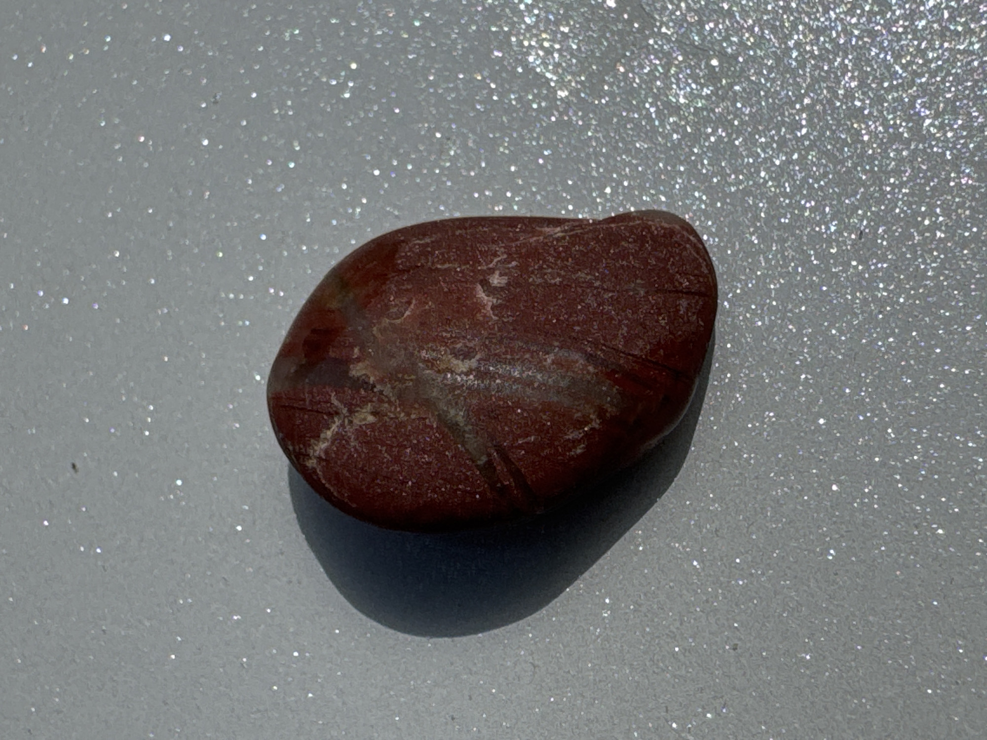 This media file displays a single, oval-shaped, reddish-brown stone resting on a surface covered in fine, reflective glitter. The stone has a rough texture and appears to have lighter-colored veins or inclusions running through it. The glittered surface is a light grey color, and the glitter particles create a shimmering effect under bright lighting. The image is a close-up shot, with the stone positioned slightly off-center, casting a subtle shadow to its right. The lighting appears to be strong, possibly sunlight, as indicated by the bright reflections on the glitter and the distinct shadow. There is no visible text in the image. The scene is static and focuses entirely on the texture and appearance of the stone and its sparkly background. The location context provided, Добровское сельское поселение, Ukraine, does not offer any visual cues within the image itself.