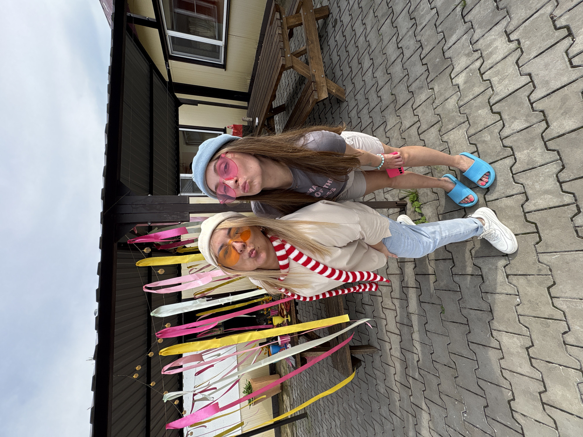 Two young women are posing for a photo outdoors in front of a covered patio area. The woman on the left is wearing a tan sweatshirt, light blue jeans, white sneakers, a white beanie, orange heart-shaped sunglasses, and a red and white striped scarf. She is leaning forward with her hands on her knees. The woman on the right is wearing a grey t-shirt, tan shorts, blue slides, a blue beret, and pink heart-shaped sunglasses. She is also leaning forward, with her right hand near her hip and her left hand holding a pink phone. The patio area is decorated with colorful ribbons and string lights. A wooden picnic table is visible behind the women. The ground is paved with interlocking concrete tiles. The scene appears to be a residential backyard or a casual outdoor gathering space. The sky is overcast.