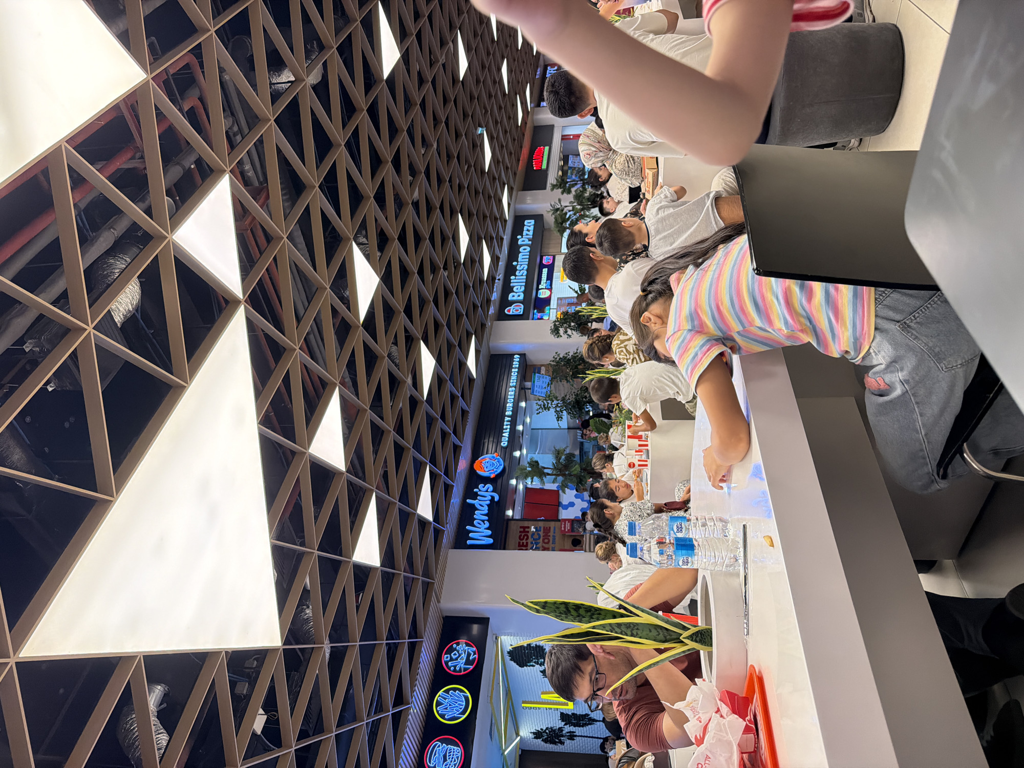 The image depicts a bustling food court likely located in a shopping mall in Tashkent, Uzbekistan. The ceiling features a modern, geometric design with a grid of wooden beams and illuminated panels. Various fast-food establishments are visible in the background, including "Wendy's" and "Bellissimo Pizza," indicated by prominent signage.
In the foreground, several people are seated at long, white tables. A man wearing glasses and a maroon t-shirt is looking to his left, with a potted snake plant on the table in front of him. Two bottles of "Bon Aqua" water are also on the table. A young girl in a colorful striped t-shirt and denim jeans is leaning her head on her arm, seemingly resting or sleeping. Other diners, including children and adults, are seen eating and interacting around the tables. The overall atmosphere suggests a busy and casual dining environment.