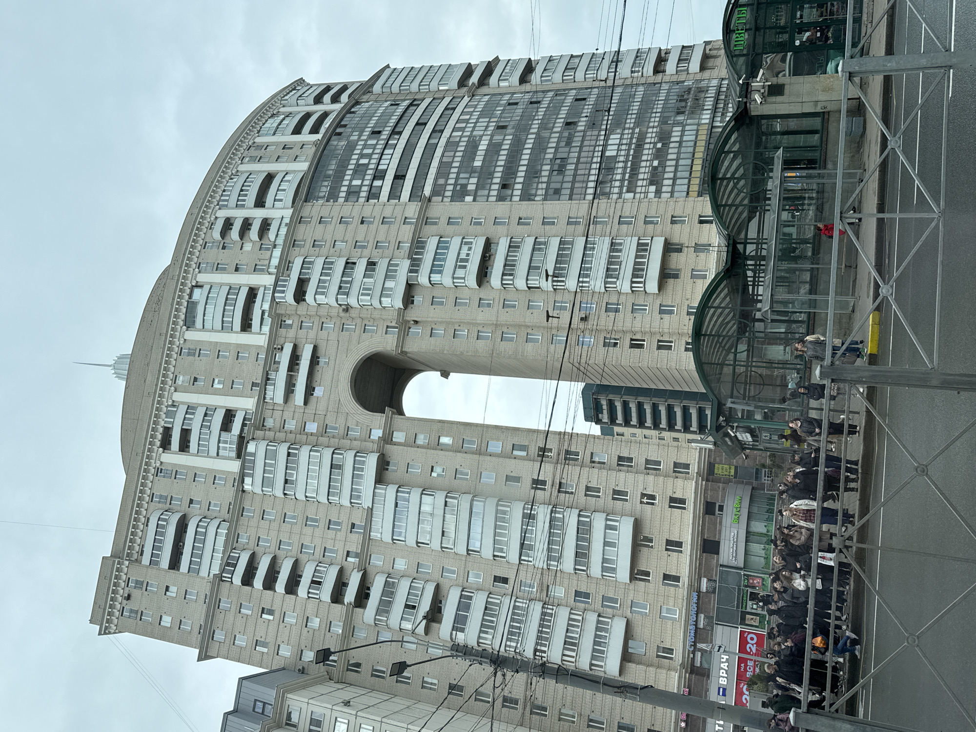 The image depicts a tall, curved residential building with a large archway in the center. This distinctive structure is located in Saint Petersburg, Russia. The building is primarily beige in color, with multiple rows of windows and protruding balconies. A smaller, more modern building is visible behind the arched opening. In the foreground, a bus stop-like structure with a green, arched roof is present. A crowd of people appears to be waiting near the bus stop. Power lines crisscross in the lower portion of the frame. The sky is overcast.