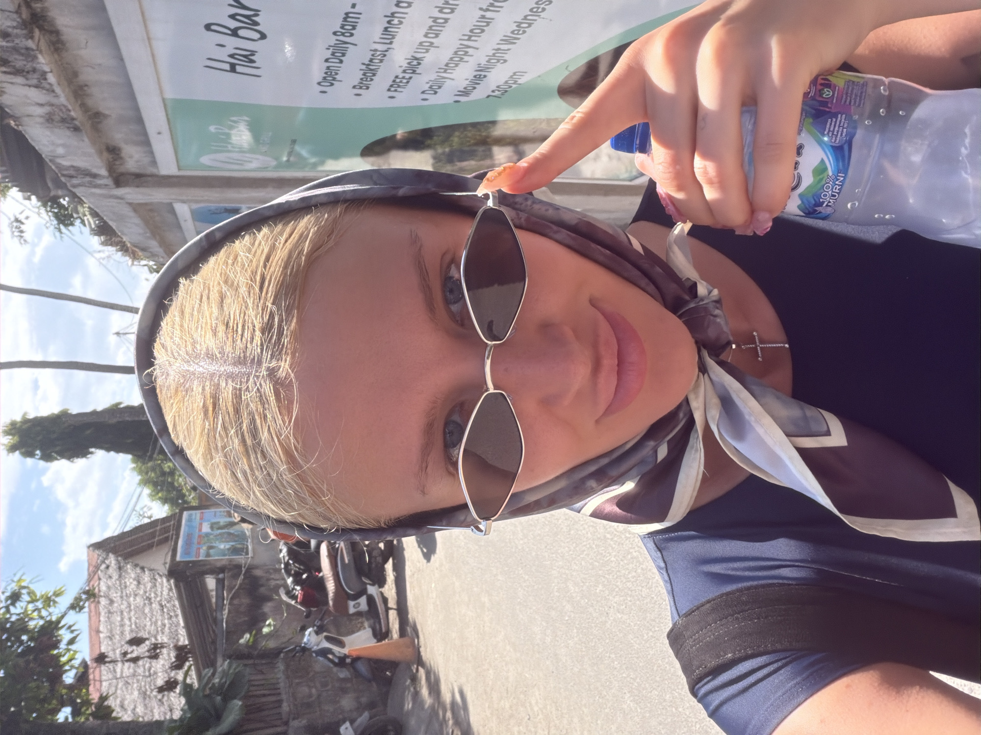 A blonde woman wearing a blue shirt, a headscarf, and diamond-shaped sunglasses is holding a bottle of water and taking a selfie outdoors in Nusa Penida, Indonesia. She is standing on a street with a concrete wall with a sign in the background. The sign lists "Hai Bar" and its opening hours and services. Behind her, there are buildings with thatched roofs and parked motorcycles. The woman is pointing her right index finger towards her sunglasses. The sun is shining, casting shadows on the ground.