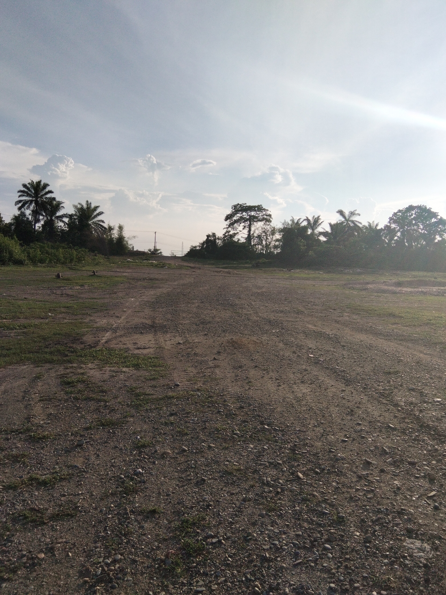 This image depicts an outdoor scene in Sukaramai Dua, Indonesia, likely during the late afternoon or early evening, as indicated by the low angle of the sun creating lens flare and long shadows. The weather appears clear with some scattered clouds in a pale blue sky.
The foreground is dominated by a rough, gravelly dirt path or road that stretches into the distance, curving slightly to the right. Patches of green grass and sparse vegetation are visible on either side of the path, suggesting an uncultivated or undeveloped area. A few small, dark wooden posts or stakes are stuck in the ground on the left side of the path.
In the background, a dense line of trees and palm trees forms a natural border. Among the trees, there are signs of human infrastructure, including utility poles with wires and what appear to be pylons in the distance, hinting at an area that is either on the outskirts of a town or in a more rural, but serviced, location.
The overall mood of the image is one of tranquility and openness, with the natural landscape taking center stage. There is no visible text in the image.