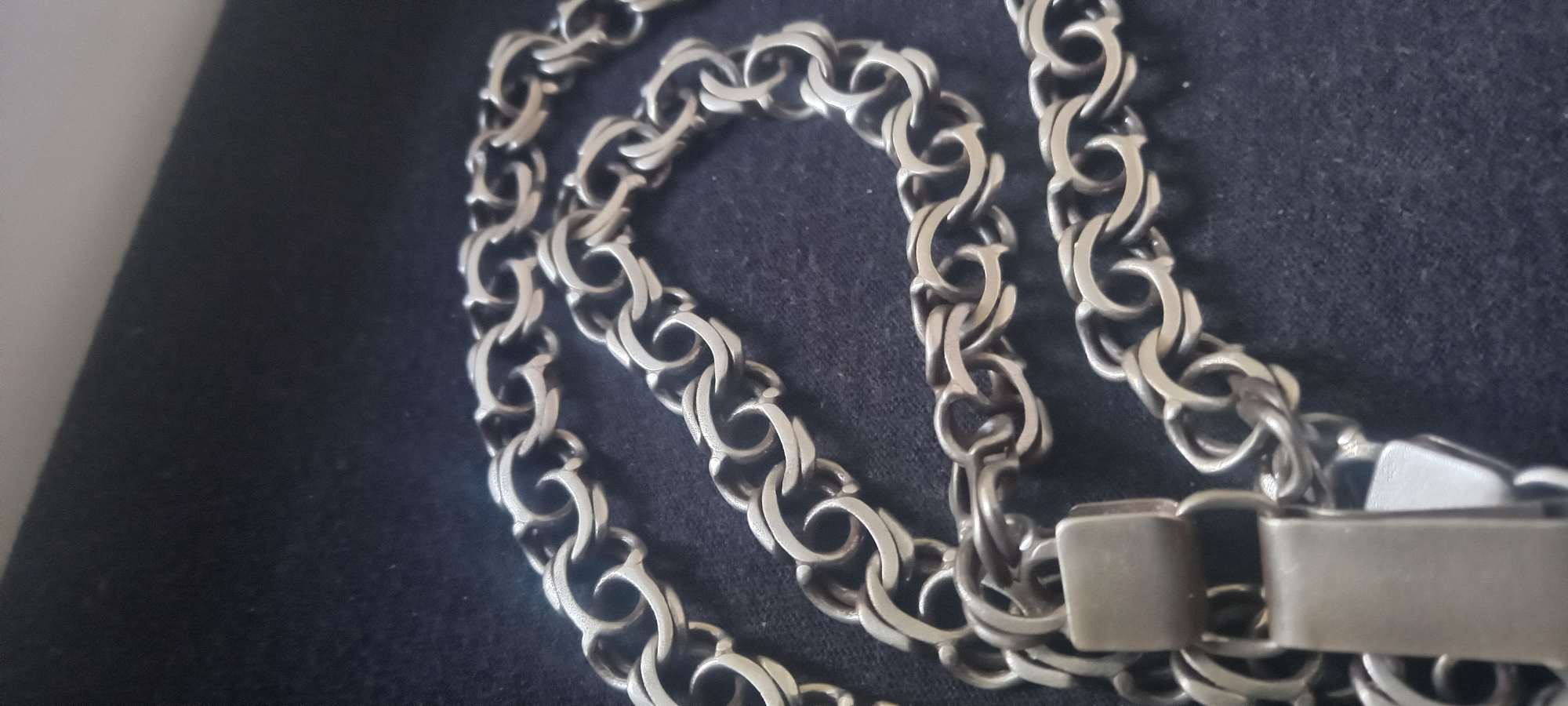A close-up shot depicts a silver chain with an intricate interlocking link design lying on a dark, textured surface. The chain includes a clasp with a rectangular design. The background appears to be out of focus. The location of the chain within the city of Kryvyi Rih, Ukraine is not identifiable from the image.