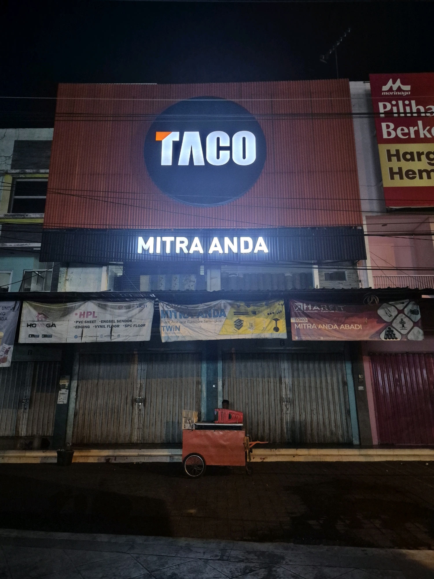 This is a nighttime photograph of a storefront in Surakarta, Indonesia. The main subject is a business with a prominent illuminated sign reading "TACO" in large white letters with an orange accent on a circular black background. Below this, another illuminated sign reads "MITRA ANDA" in white letters on a black background. The storefront is covered by corrugated metal roll-up doors, which are mostly closed. A bright orange food cart with two wheels is parked in front of the closed doors, occupied by a person wearing a dark shirt. Banners hang above the roll-up doors; one banner on the left displays "HOMEGA", "HPL", "PVC SHEET", "ENGSEL SENDOK", "EDGING", "VYNIL FLOOR", and "SPC FLOOR". Another banner in the center reads "MITRA ANDA TWIN" and appears to be advertising furniture. A third banner on the right advertises "HARFIT HARDWARE - FURNITURE FITNESS" and "MITRA ANDA ABADI". Other smaller signs and logos are visible on these banners and the building facade. The street in front of the storefront is paved with interlocking bricks and appears wet, reflecting the lights. Electrical wires are strung across the upper portion of the image. The overall lighting suggests it is after dark, with artificial illumination from the signs and a faint ambient light.