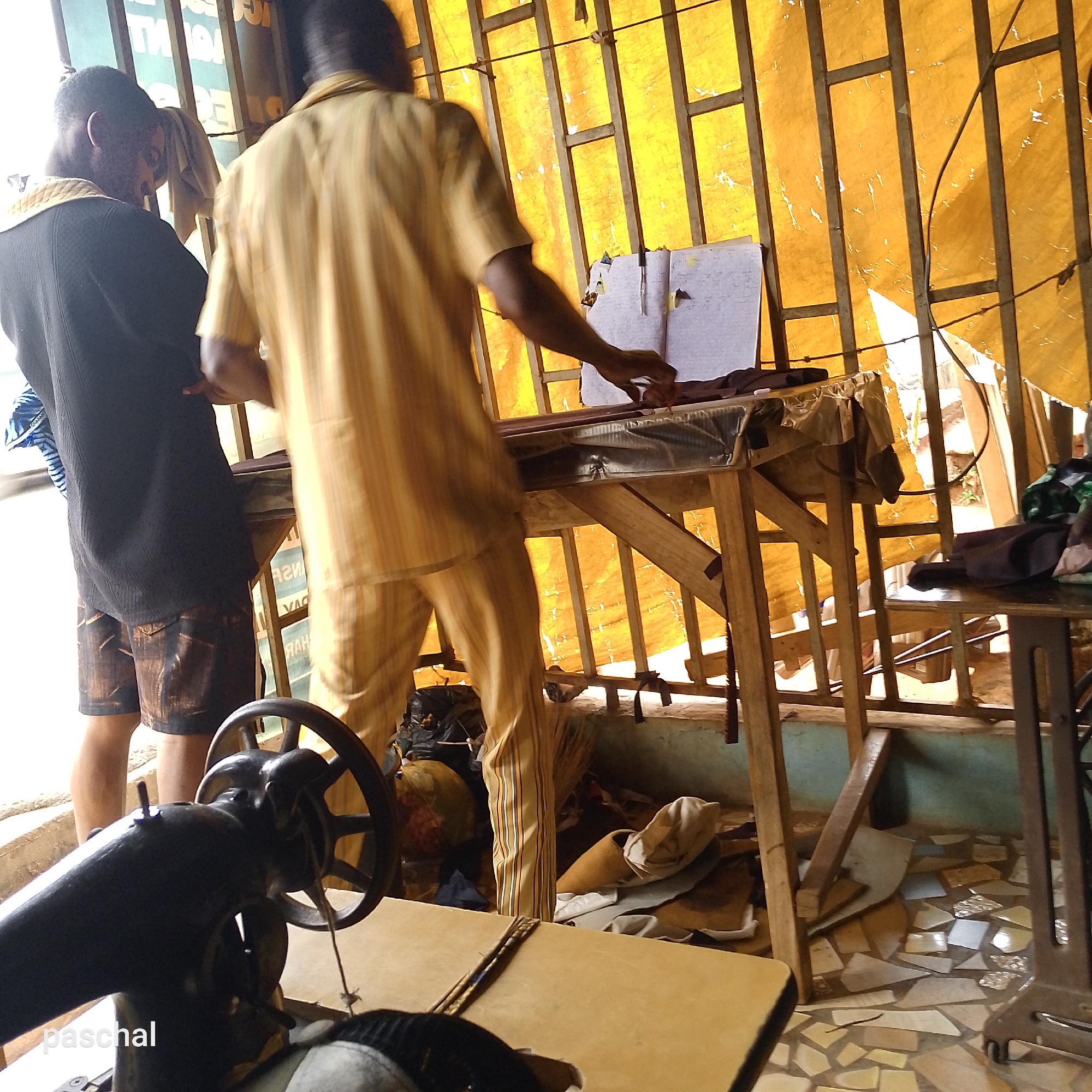Nsugbe, Nigeria. Two men are standing near a table with an open notebook. One man is wearing a black sweater and shorts. The other is wearing a striped yellow shirt and pants. The men appear to be tailors, with a sewing machine visible in the foreground. The environment is cluttered with cloth scraps and other items related to tailoring. A yellow curtain hangs in the background. The floor is decorated with broken tiles.