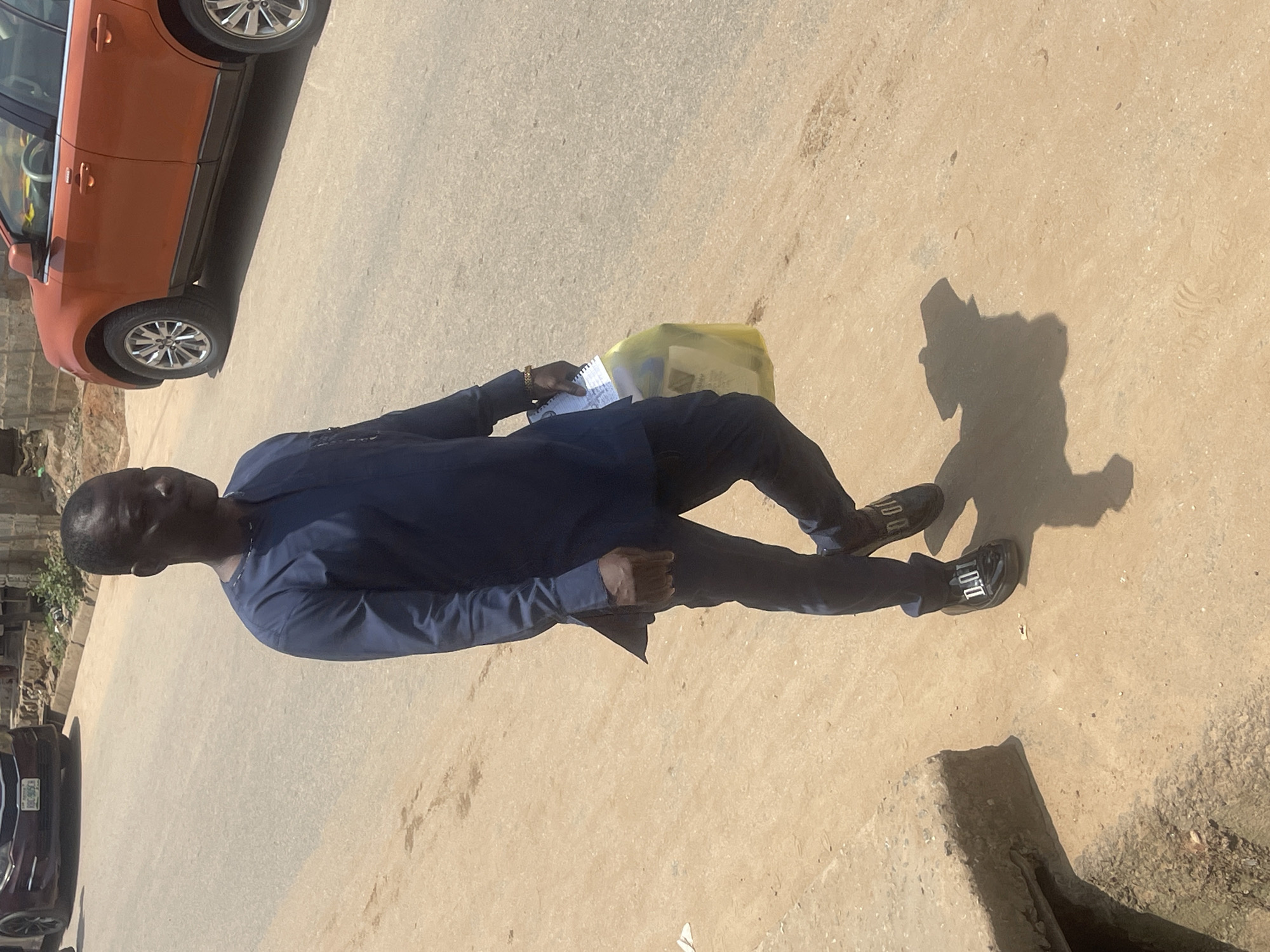 The image shows a man in a dark blue traditional outfit standing on a paved surface, likely a road or sidewalk. He is holding a notepad and pen in his right hand, and a yellow plastic bag is visible next to his left leg. The man is wearing dark shoes with white lettering that reads "LOUI". A portion of an orange car is visible in the upper left corner of the image, and a dark-colored car with a license plate is partially visible in the lower left. The scene appears to be outdoors during the day, with sunlight casting a shadow of the man on the ground. The setting suggests an urban or semi-urban environment in Abuja, Nigeria. There are no clear indications of the weather or the man's emotions, but the overall impression is that of a casual moment. The visible text on the shoes is "LOUI".