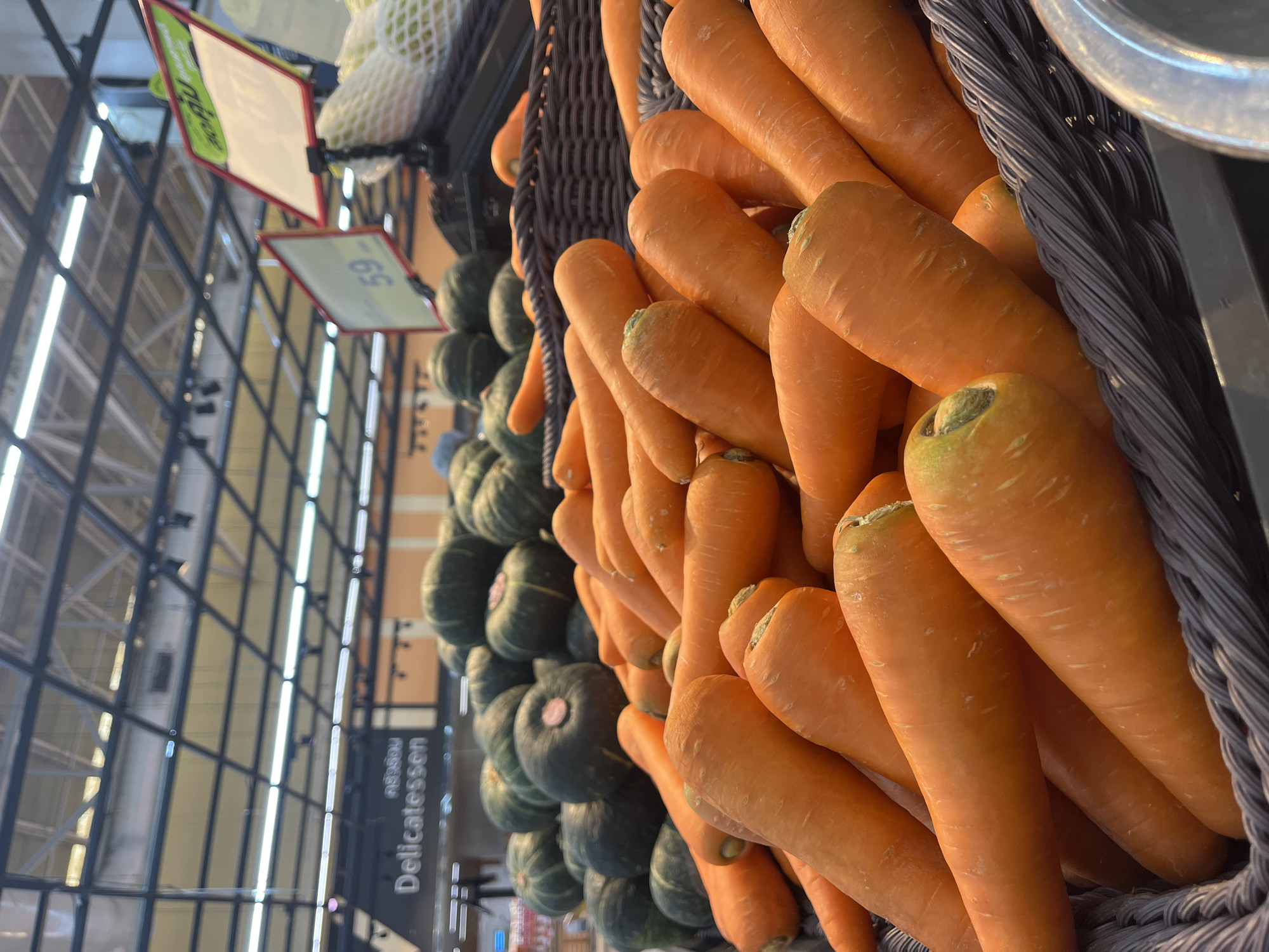 The image shows a close-up view of a basket filled with fresh carrots in what appears to be a grocery store or market. The carrots are bright orange and have a slightly tapered shape, with visible root ends. Behind the carrots, there are more vegetables, including several round, dark green squash or pumpkins, also piled in a display. In the background, overhead lighting fixtures and store shelving are visible, along with a sign that reads "Delicatessen". There are also price tags visible, one of which displays the number "59". The lighting suggests an indoor setting, likely a commercial establishment. The scene depicts a produce section of a store.