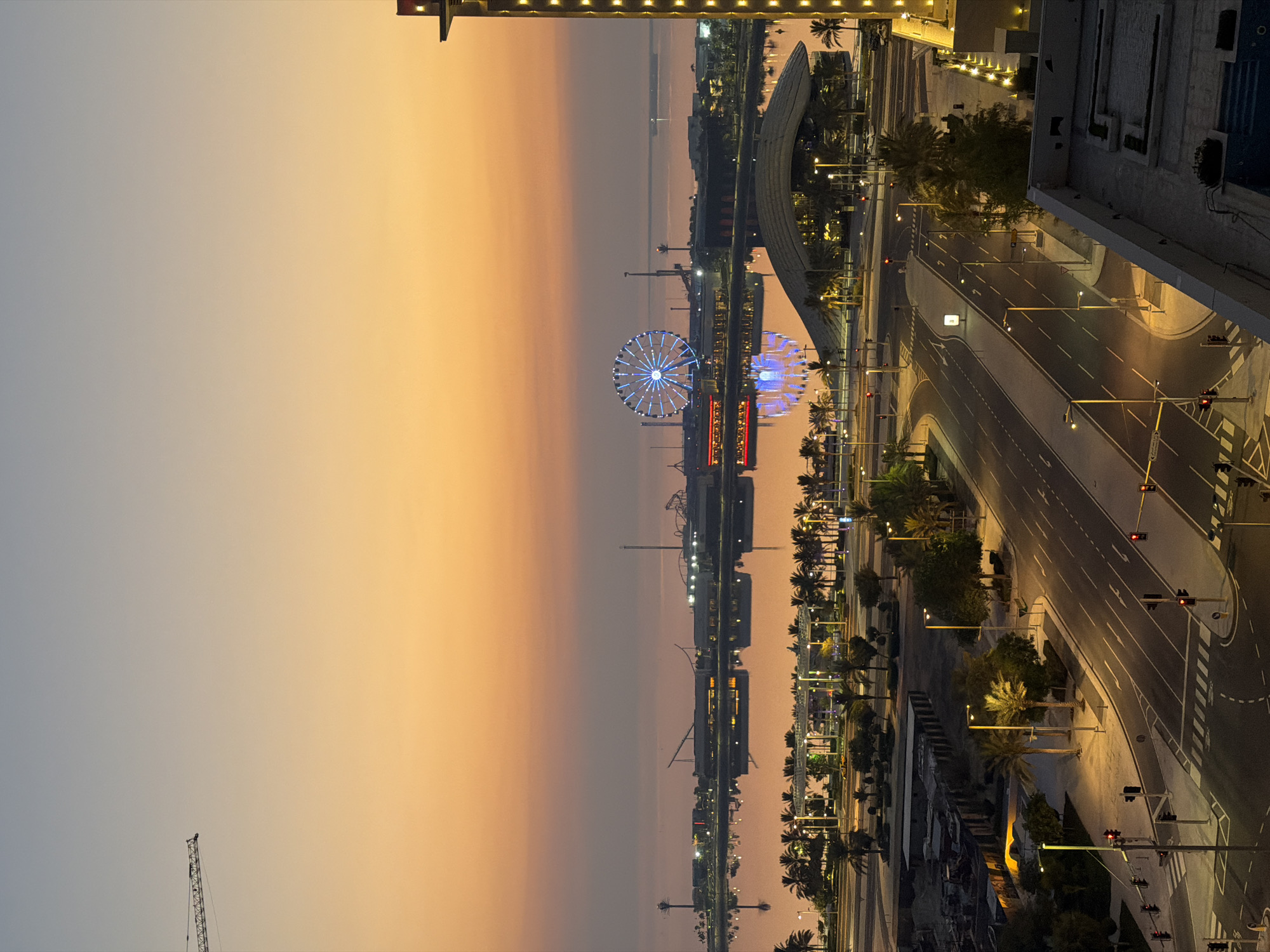 The image depicts a wide, elevated view of a city skyline at dusk. The sky transitions from a pale blue at the top to a warm orange and yellow gradient towards the horizon, suggesting the end of daylight. In the distance, a large Ferris wheel illuminated with blue lights stands prominently. Adjacent to the Ferris wheel, a collection of amusement park rides and buildings are visible, with lights reflecting on a body of water in front of them. A modern, curved architectural structure with glowing lights is situated closer to the foreground. The middle ground features a multi-lane road with traffic lights and street lamps, lined with palm trees and some construction activity. The foreground shows the rooftop of a building, with what appears to be a swimming pool and some planters. A construction crane is visible in the upper left corner against the pale sky. No people are clearly visible in the image. The scene conveys a sense of urban development and leisure, possibly a waterfront entertainment district.