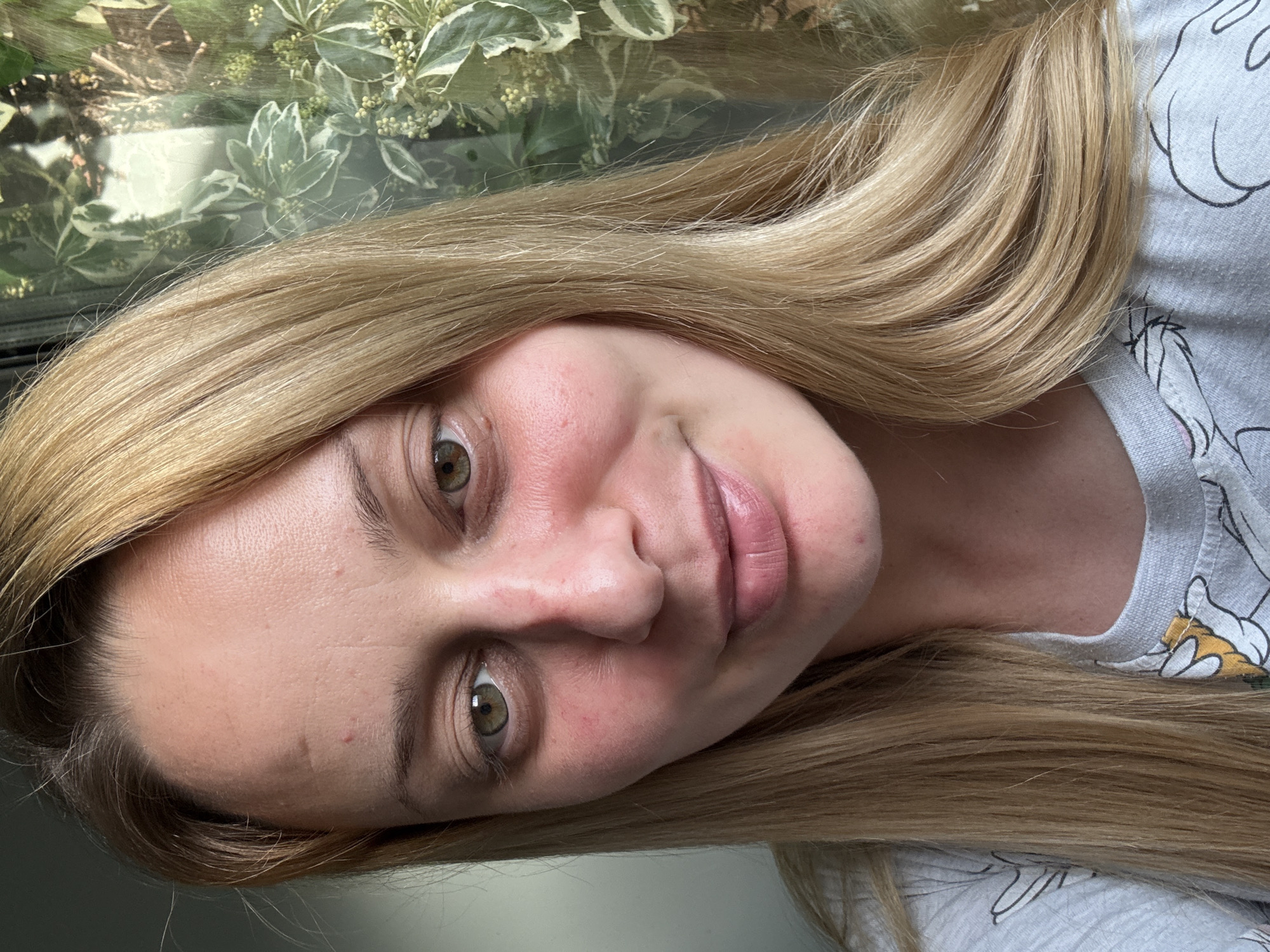 A woman with light blonde hair, styled with a side part and long, loose waves, is facing the camera. She has fair skin, green eyes, and pinkish lips. There are slight skin imperfections on her face. She is wearing a light gray shirt with a cartoon character design. Behind her is greenery, potentially plants in a window. There is no definitive evidence to indicate a location in Hazlemere, United Kingdom, or any other specific location.
