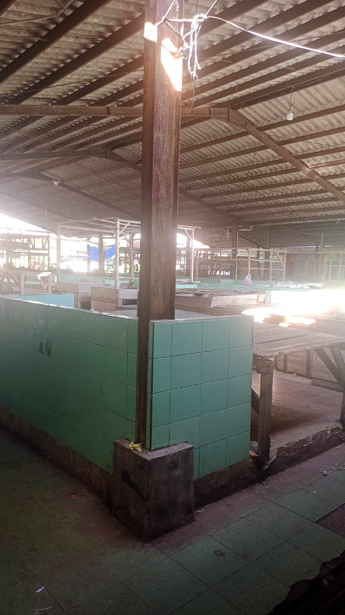 The image depicts an empty market stall in Cogreg, Indonesia, under a corrugated metal roof supported by wooden beams and a central concrete pillar. The stall features turquoise tiled walls and a concrete base. Beyond the stall, other market stalls, some with wooden tables and structures, are visible in the background, with evidence of sunlight filtering through the roof. The ground is tiled with green squares. The scene suggests the market is either closed or has not yet opened for the day, as no people are present.