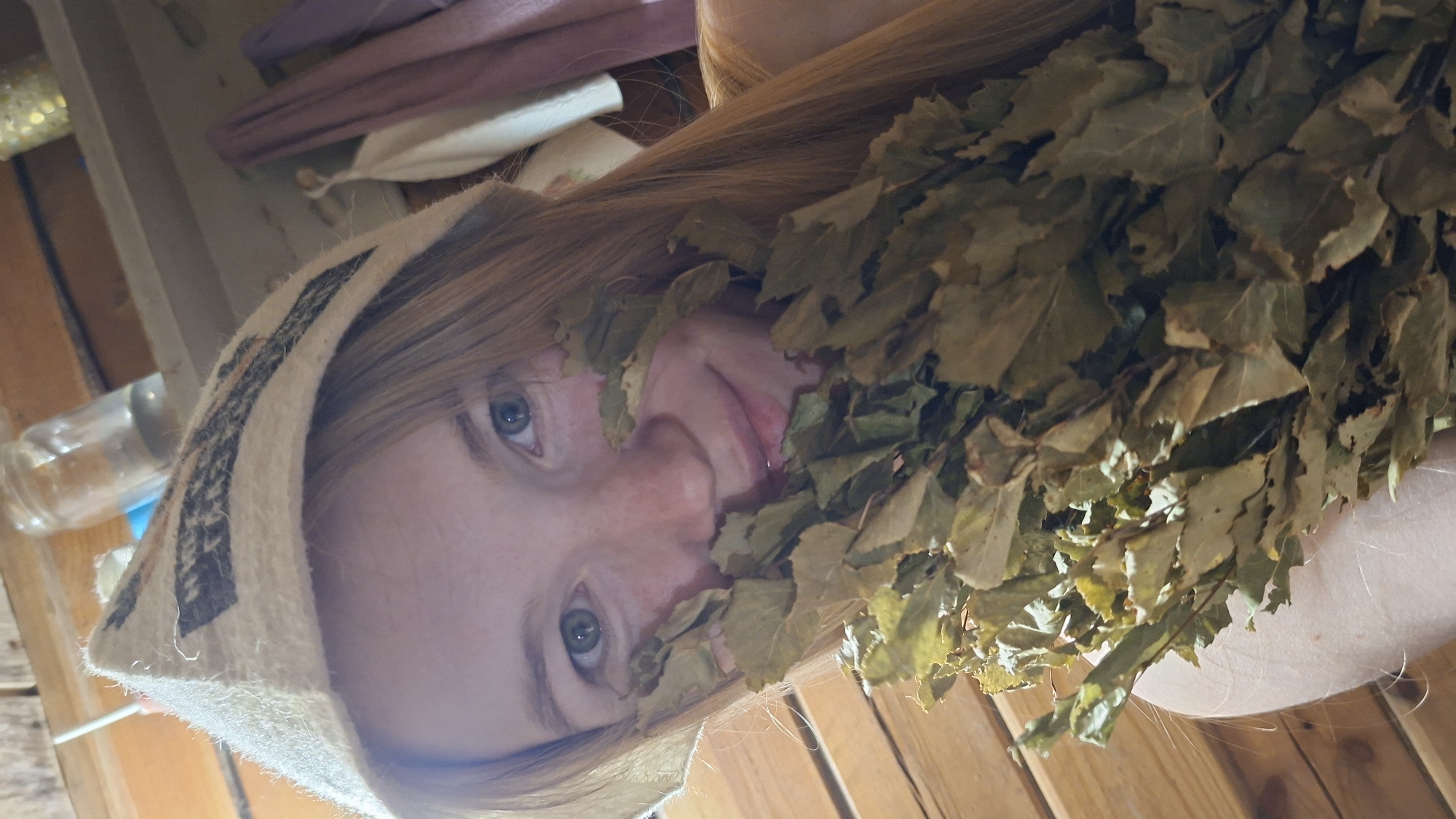 A young woman with long red hair and green eyes, wearing a white felt hat with a patterned band, is partially obscured by a bundle of dried leaves, possibly a venik for a banya. She is in what appears to be a banya or sauna, indicated by the wooden plank walls. The background shows shelves with a glass jar containing a clear liquid and other blurred items. The lighting is warm and natural, suggesting an indoor setting. The scene is a close-up, capturing the woman's face and the leaves. The location is identified as Bolshoy Bukor, Russia.