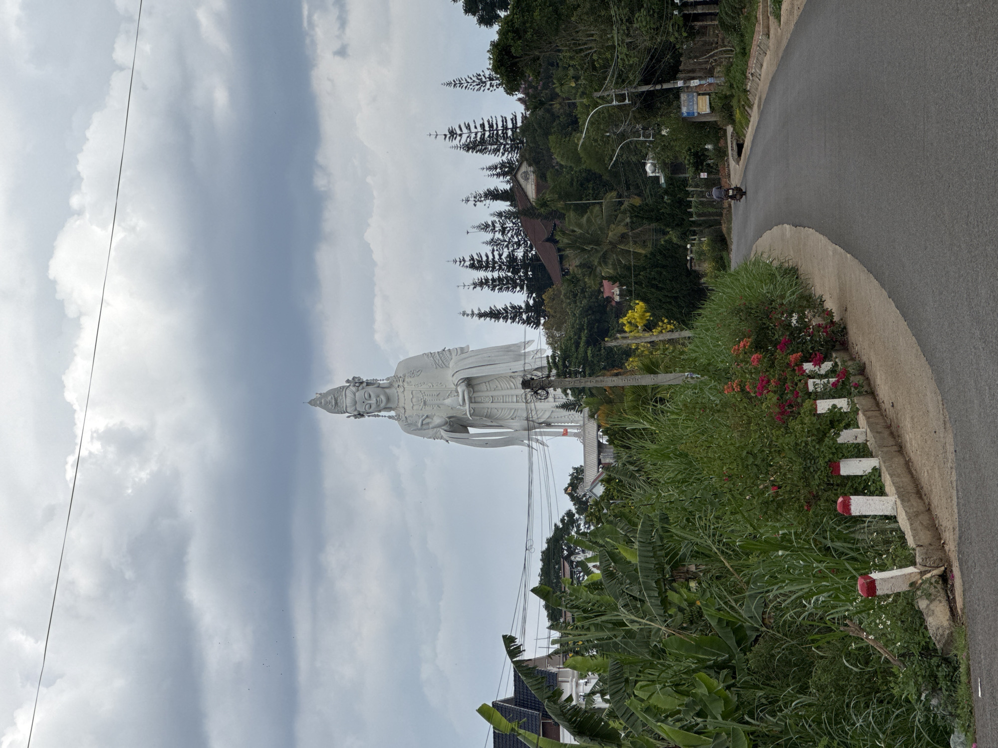 The image shows a tall, white statue of a woman in Nam Ban, Vietnam, standing adjacent to a road. A motorcyclist is riding on the road. Lush greenery and trees surround the statue and line the roadside. The sky is overcast with cloudy weather. A fence is visible along the road edge. Houses are visible at the bottom of the image. Power lines are running over head.