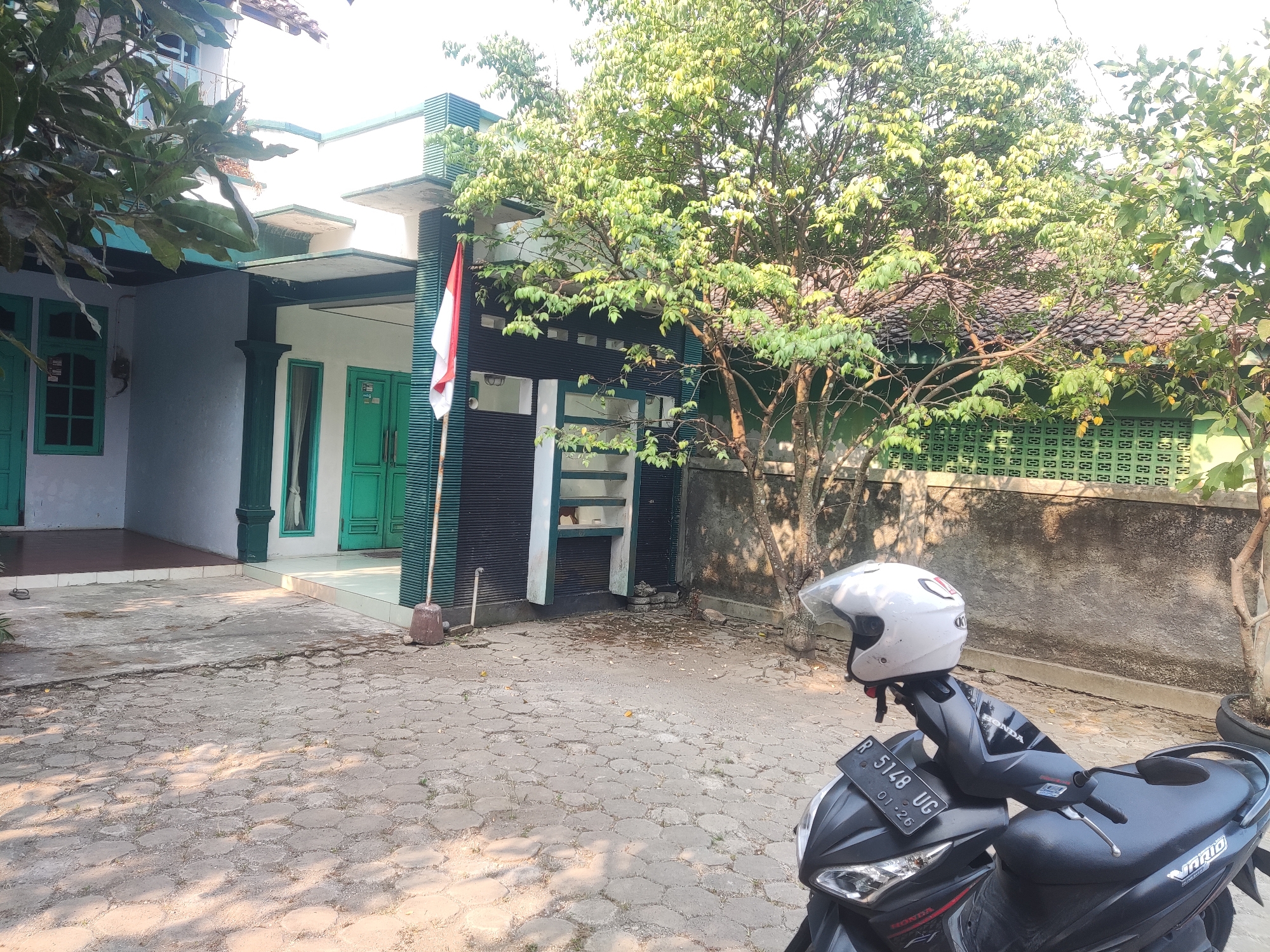 The image depicts a street scene in Godean, Indonesia, on a sunny day. In the foreground, a black motorcycle is parked, equipped with a white helmet resting on its handlebars. The motorcycle's license plate reads "R 5148 UG 01-26". Adjacent to the motorcycle is a broom leaning against a wall.

The background features a house with bright green doors and windows, characteristic of Indonesian architecture. A flagpole with the Indonesian flag stands to the left of the house. Trees with lush green foliage are prominent, casting dappled shadows on the paved ground. The overall environment suggests a residential area in a tropical climate.
