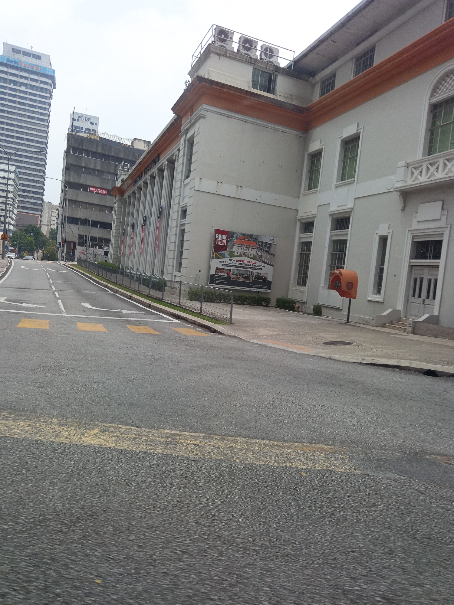 The image captures a street scene in Kuala Lumpur, Malaysia, featuring a prominent building with classical architectural elements. The building, painted in a light gray with orange trim, has a colonnade along its facade and large, tall windows. A large banner is affixed to the building's exterior, advertising "New Event Space Available at Muzium Telekom." To the left of this building, a narrower, darker building stands, and further in the background, a tall, modern skyscraper with a blue top is visible. The street in the foreground is asphalt, marked with yellow pedestrian crossing lines and white lane dividers. Several cars are seen on the road in the distance, and there is a traffic light visible. A distinctive orange, booth-like structure is positioned on the sidewalk in front of the gray building. The sky is overcast, suggesting a cloudy day.