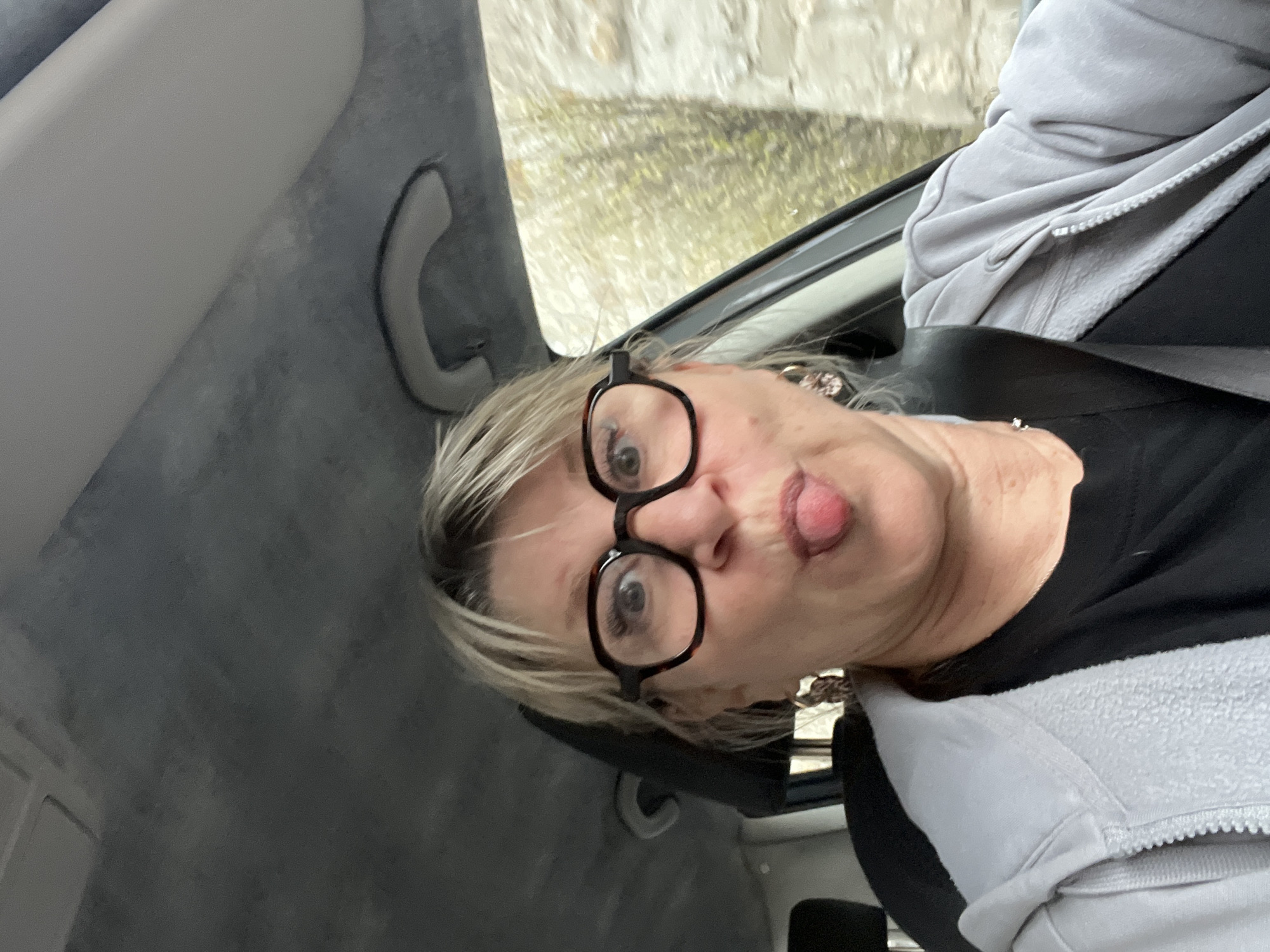 A woman is in the passenger seat of a car, wearing black framed glasses. She has blonde hair and is wearing a black shirt under a grey zippered jacket. She is sticking out her tongue and making a funny face. The interior of the car is grey. Through the window, a blurred view of the outside is visible, appearing to show a stone wall and greenery. It is unknown if the location is in Saint-Galmier, France.