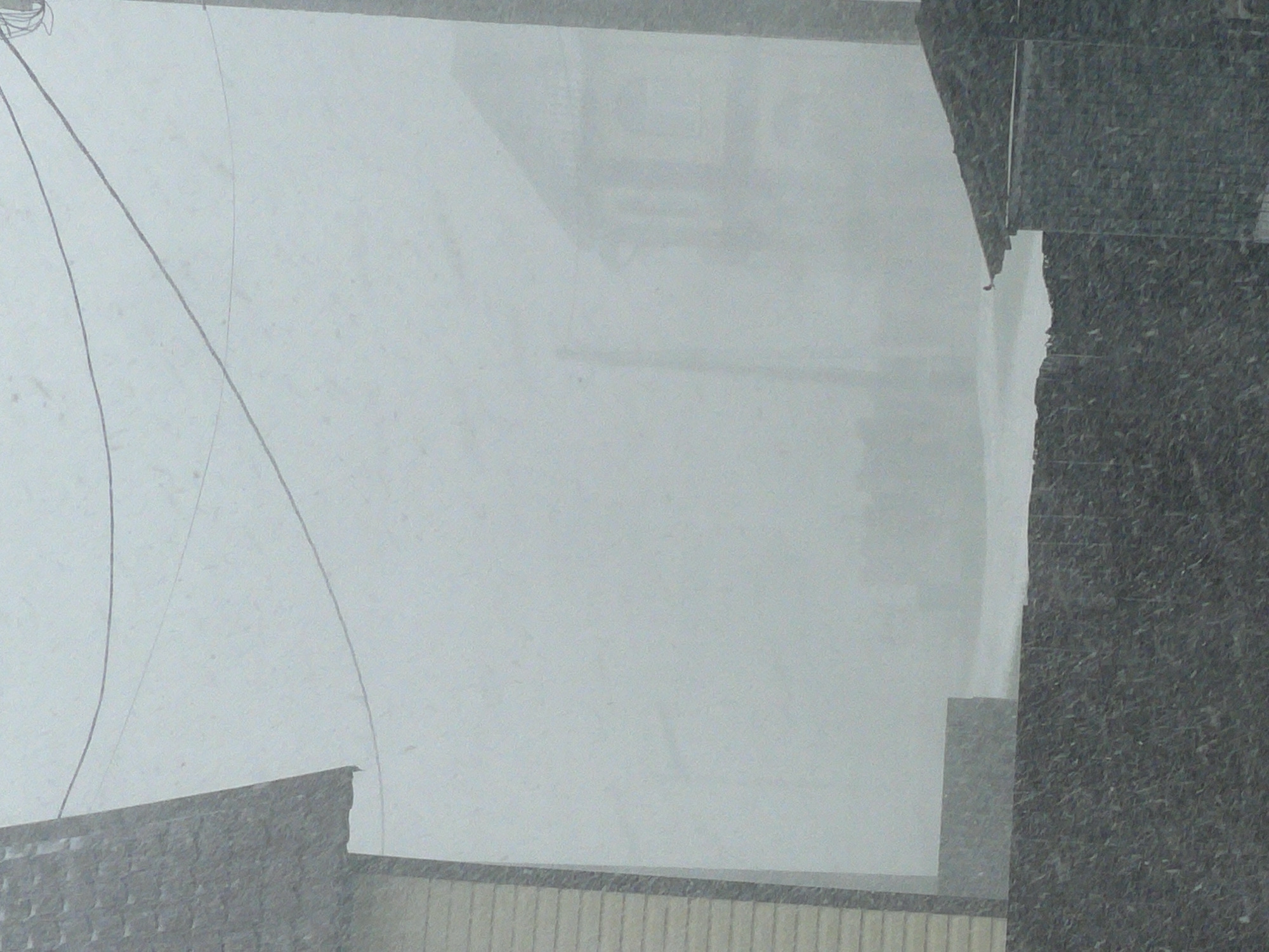 The photograph appears to be taken from inside a building, looking out onto a snowy cityscape in Chulym, Russia. The view is partially obscured by a window and the edge of the building's structure, made of a dark, textured material. In the distance, the outlines of other buildings are visible through the snow-filled air, but the details are indistinct due to the heavy snowfall. Wires extend downwards into view. The scene suggests a cold, overcast day in an urban environment, likely taken from an elevated position.