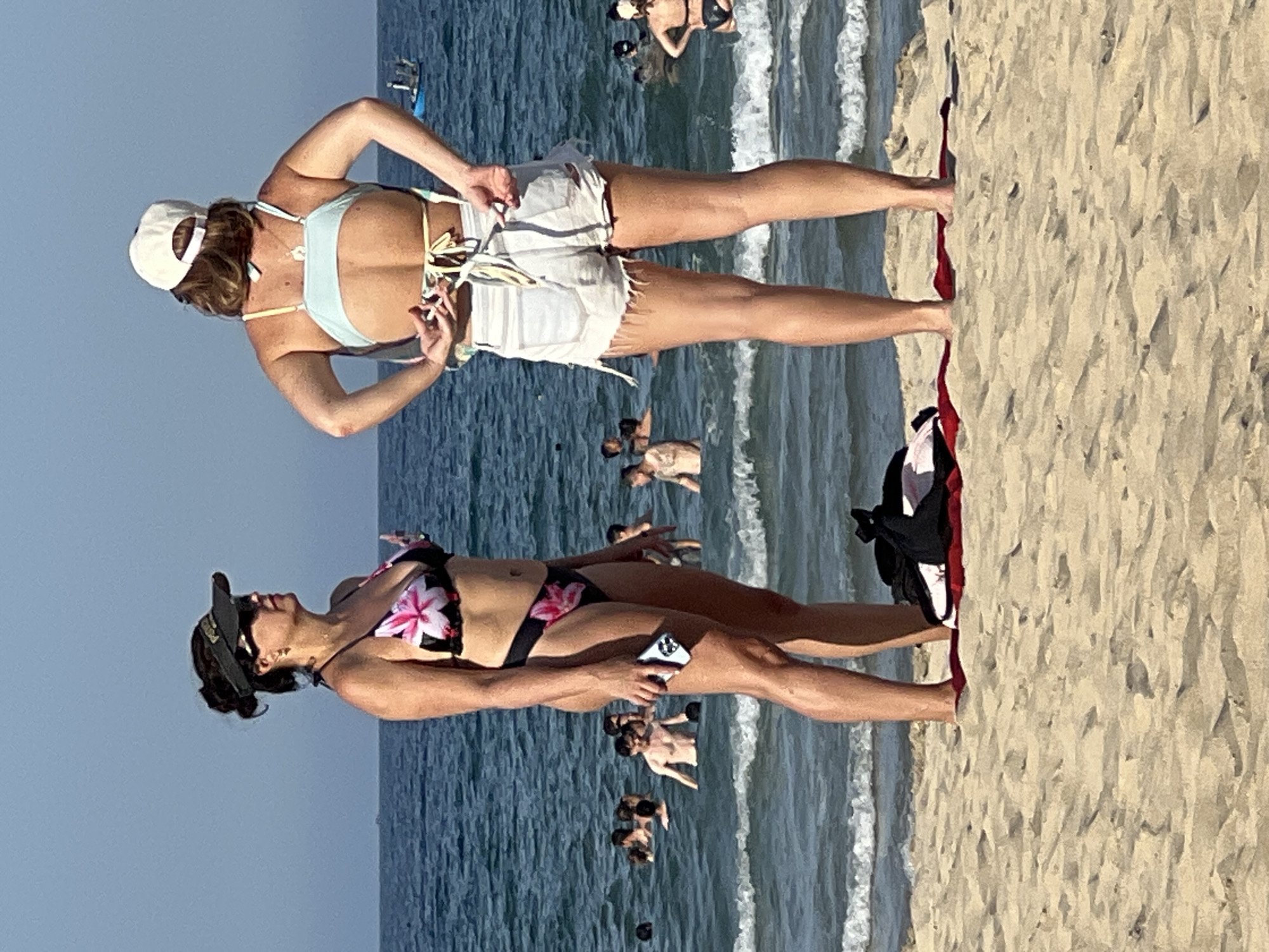 Two women are on a beach in Đà Nẵng, Vietnam, with the ocean and other beachgoers in the background. The woman on the left is wearing a black and pink floral bikini, a dark visor, and sunglasses. She is holding a cell phone in her right hand and looking towards the ocean. The woman on the right is wearing a light blue bikini top and distressed white denim shorts. She has a white baseball cap on backwards and is adjusting the tie on her bikini top. There are several people swimming and wading in the ocean. The sand is light brown, and there are waves breaking near the shore. A red towel and a black bag are on the sand in front of the women. The sky is clear and blue.