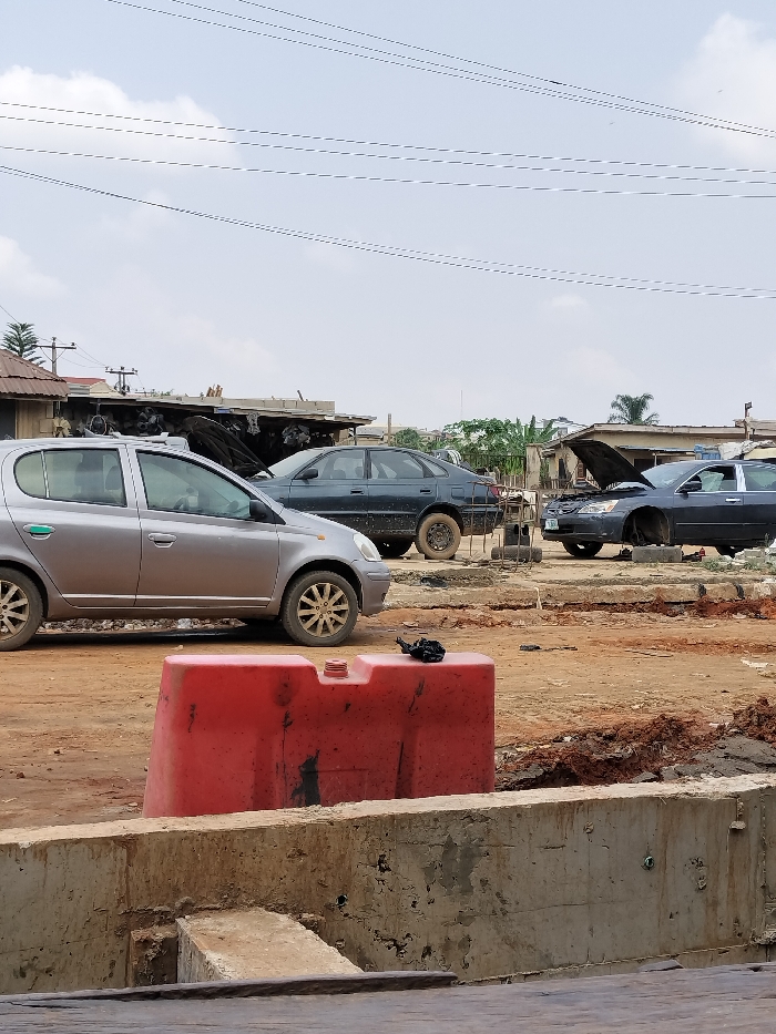 The image depicts an outdoor area in Magboro, Nigeria, characterized by vehicles and structures on an unpaved, reddish-brown dirt surface beneath an overcast sky.
In the immediate foreground, a red plastic road barrier, consistent with a water-filled barrier, is centrally positioned on a concrete base. A red cap is visible on the barrier's top surface, alongside a small, dark, crumpled object.
Behind the barrier, a silver five-door hatchback vehicle, identified as a Toyota Yaris/Vitz model, with mud-splashed wheels, is parked facing right. To its rear-right, a dark blue sedan, consistent with a Peugeot 406, is parked, appearing to be missing its front driver-side wheel and resting directly on the ground. Further to the right, another dark sedan is parked with its hood open, indicating a state of repair or disassembly. This vehicle also appears to be missing its front driver-side wheel, resting on unseen supports or the ground. The midground consists of uneven, reddish-brown earth with visible ruts.
In the background, several single-story structures, likely workshops or storage areas, with corrugated roofs, are visible. These structures are interspersed with sparse green vegetation, including a palm tree. Multiple utility lines stretch horizontally across the pale, cloudy sky. A utility pole with transformers is visible on the far left. No individuals are present in the scene.