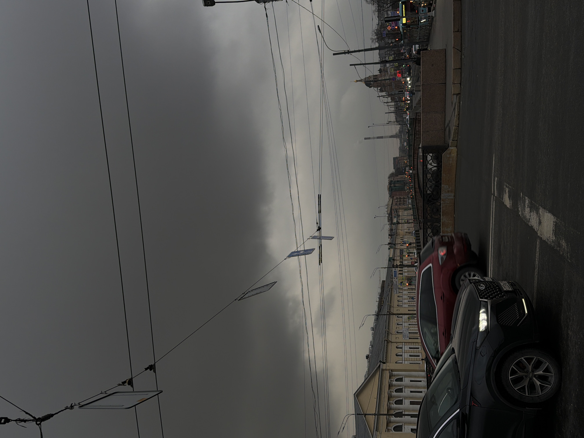 The image depicts a street scene in Saint Petersburg, Russia, taken from a low angle. The sky is overcast and dark. A street with at least two cars, a dark SUV in the foreground and a red car behind, is visible on the right. The street appears wet, reflecting light. Buildings with architectural details line the street, with windows and cornices visible. Overhead wires and power lines crisscross the sky. In the distance, a skyline with various structures, including possibly a church spire and industrial elements, is visible. The overall impression is one of a cloudy, urban landscape in Saint Petersburg.