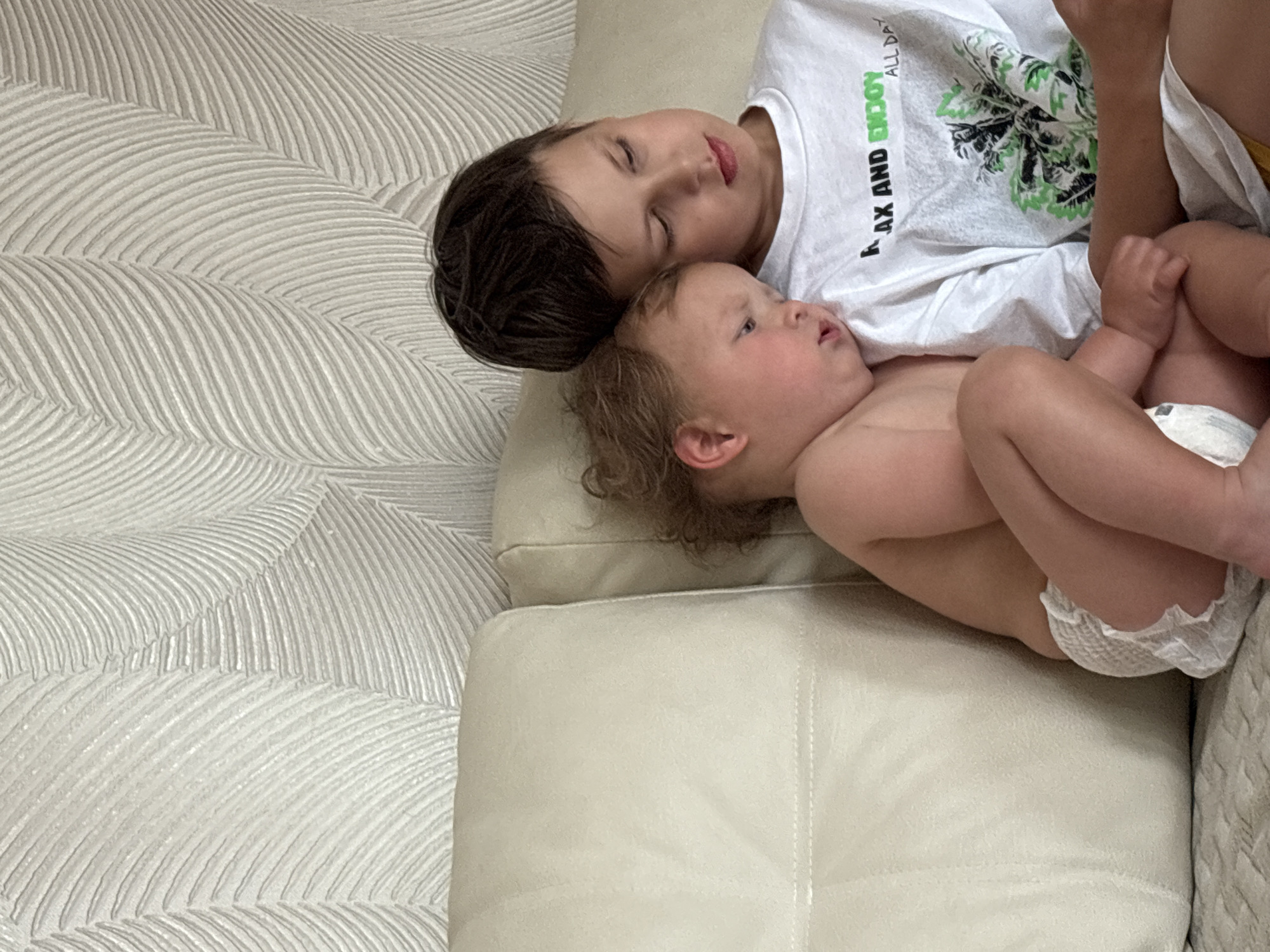 Two children, an older boy and a baby, are lying down on a cream-colored sofa. The older boy has his eyes closed and his head is resting on the baby's head. He is wearing a white t-shirt with green text that reads "RELAX AND ENJOY ALL DAY" and an image of palm leaves. The baby, wearing only a diaper, is looking to the side with their mouth slightly open. The sofa is placed against a wall with textured wallpaper featuring a pattern of wavy lines. The scene appears to be indoors, likely in a home, with soft lighting suggesting it could be daytime. The overall mood is peaceful and relaxed, with the children resting together.