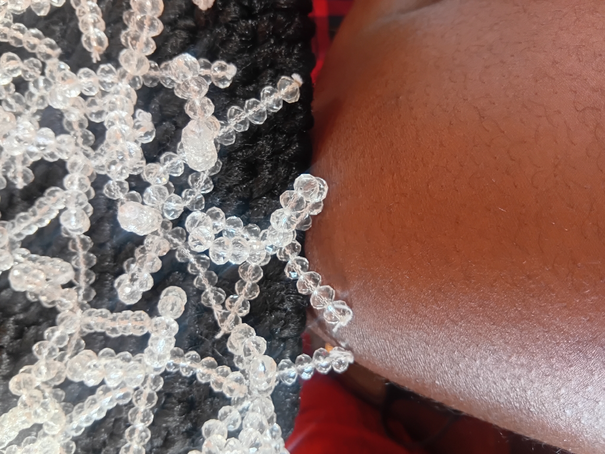 The image is a close-up shot of a person's leg and a beaded accessory. The skin appears to be brown and slightly textured, with some visible pores and fine hairs. The accessory is made of clear, faceted beads strung together to form a delicate pattern. The beads are reflective, catching the light and creating a shimmering effect. The accessory is resting on a dark, textured surface, possibly fabric. A hint of red and black plaid fabric is visible in the background on the left side of the image. The overall impression is one of a personal adornment or decoration. There is no discernible location in the city of Ejule, Nigeria within the image itself.