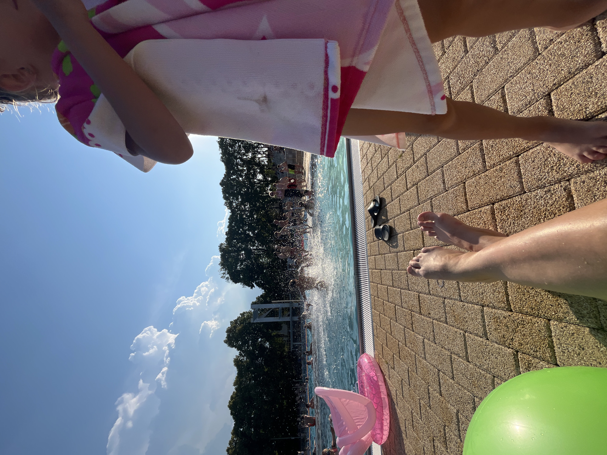 An outdoor swimming pool area on a sunny day, with a blue sky and fluffy white clouds. In the foreground, a person's legs and feet are visible, with a green inflatable ball to the left. A pink inflatable swan float rests on the tiled pool deck. In the middle ground, people are swimming and splashing in the pool. In the background, a line of trees borders the pool area. A young girl in a pink and white towel is in the right foreground, partially obscuring the view of the pool.