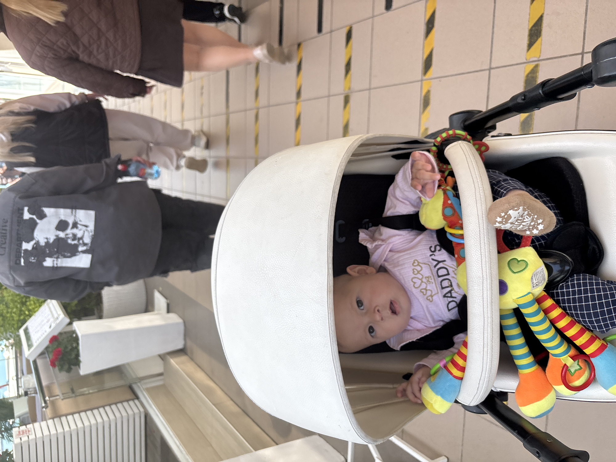 A baby in a white stroller looks up at the camera with an open mouth. The baby is wearing a white shirt with the word "Daddy's" on it, surrounded by hearts. A colorful toy hangs from the stroller's bar, near the baby. Outside the stroller, pedestrians walk along a sidewalk paved with tiles that have yellow and black stripes. In the background are commercial buildings, one of which has potted red flowers. The location is determined as Sochi, Russia.