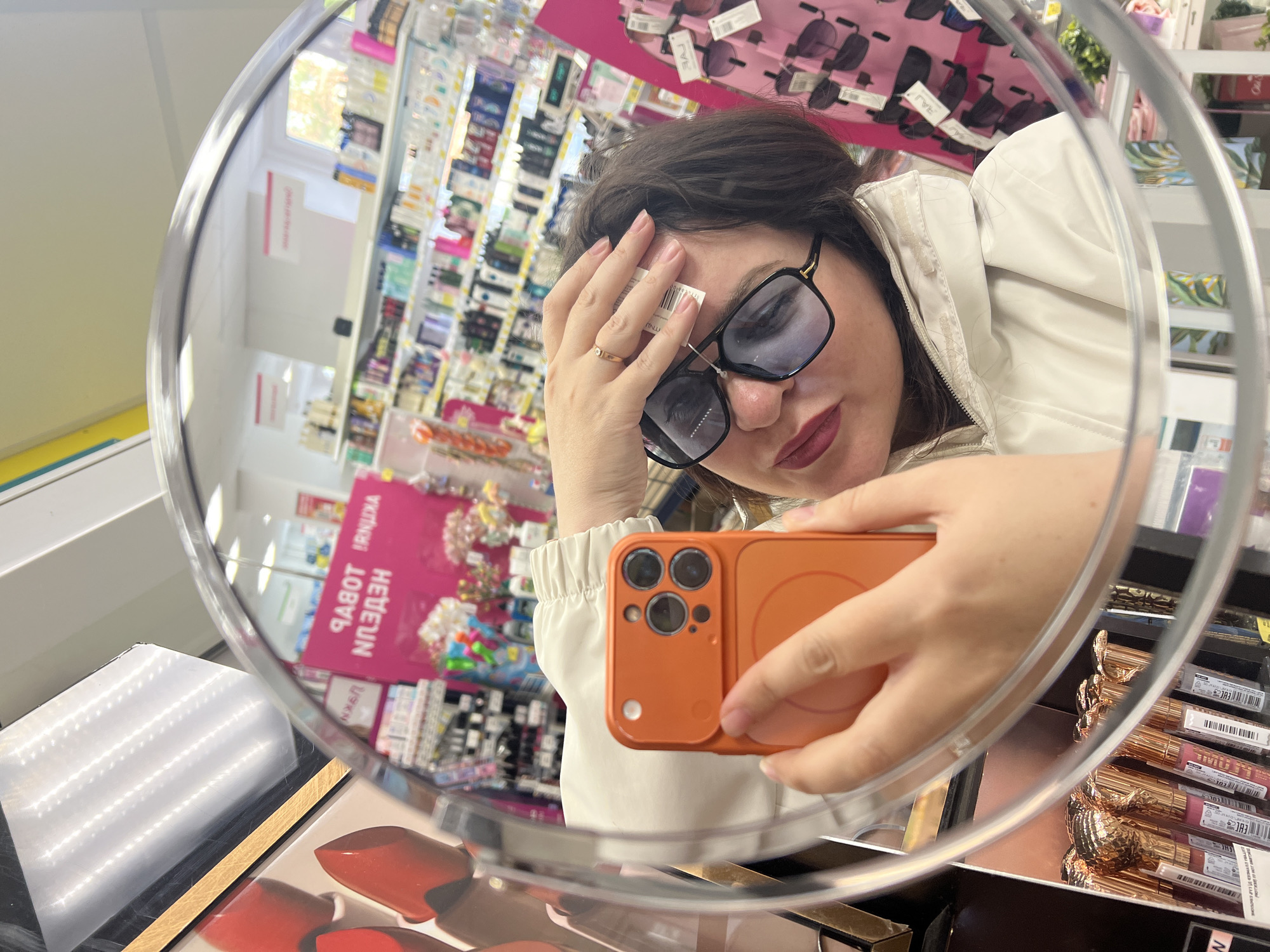 A person is taking a selfie in a mirror inside a store. The person is wearing sunglasses with a price tag still attached and a light-colored jacket. They are holding an orange smartphone. Cosmetics and other items are visible on shelves in the background and in the foreground on the shelves beneath the mirror. A sign with text in Russian, possibly "НЕДЕЛЯ РАБОТ", is visible in the background. Based on the text on the packaging of goods "Сделано в России/ Made in Russia", and the text on the sign with Russian characters, it can be assumed that the location is in Russia. The city of "Noviy" is not discernible from this image.