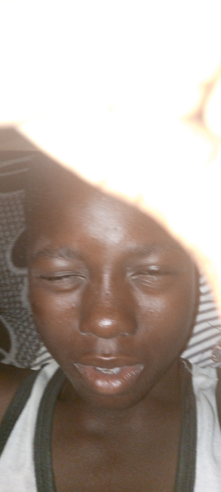 A close-up, poorly lit, selfie-style photograph shows a person with dark skin and closed eyes. The person appears to be lying down, with a patterned fabric visible to the left and striped fabric visible to the right. The person is wearing a white tank top with dark trim around the armholes. Part of a hand and bright light source is obscuring the top portion of the image. The location is unknown, and there is no indication that it is located in Adankolo, Nigeria.