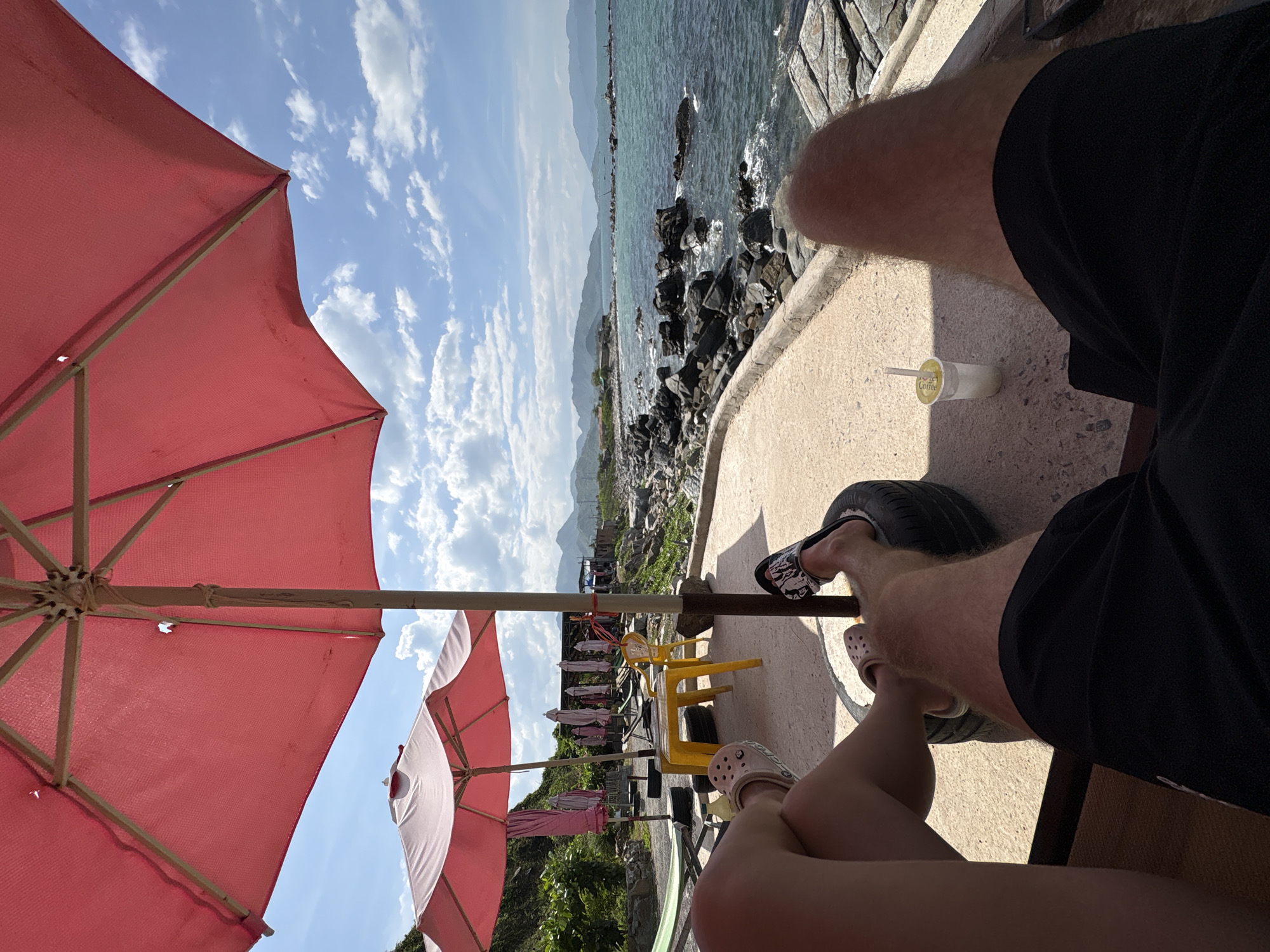 An overhead shot captures two individuals enjoying a seaside view in Vĩnh Lương, Vietnam, seated under a large pink umbrella. One person, wearing pink Crocs, has their legs crossed while resting on a repurposed tire. The other, wearing black and white patterned slides, also has their feet on a tire, with black shorts visible. In the background, a turquoise sea meets a rocky coastline under a partly cloudy sky. A small yellow table and chairs are set up nearby, along with other folded umbrellas and lounge chairs, suggesting a beachside establishment. A cup with a straw sits on the concrete ground next to the seated individuals, hinting at a refreshing drink.