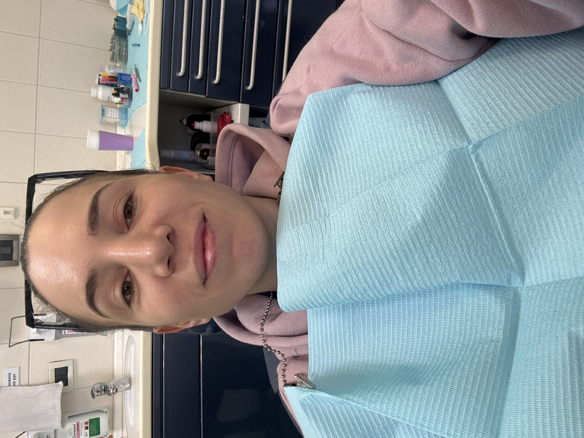A young woman is lying in a dental chair, covered with a light blue bib. She is wearing a pink hooded sweatshirt. The dental office has a set of drawers and a sink with soap and paper towels nearby. The location is not in Moscow, Russia; the sign is in Ukrainian.