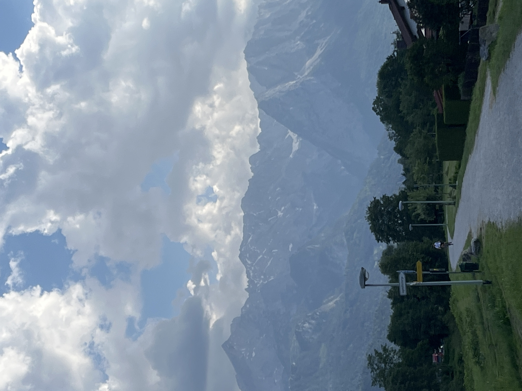 This media file depicts a scenic view in Garmisch-Partenkirchen, Germany. The foreground features a paved pathway bordered by lush green grass and trees. Several streetlights and directional signs are visible along the pathway. One of the signs has text in a foreign language, likely German, but it is not legible. In the middle ground, a person is riding a bicycle along the path. The background is dominated by the imposing presence of a mountain range, partially obscured by clouds. The mountain slopes show patches of snow, suggesting a higher altitude. The sky is a mix of white, fluffy clouds and patches of blue, indicating possibly overcast or partly sunny weather. The overall impression is a serene, outdoor setting, likely in the late afternoon or early evening given the soft light and the muted colors. The combination of the cultivated pathway, the natural landscape of mountains and greenery, and the presence of human activity (cycling) creates a harmonious and inviting scene.