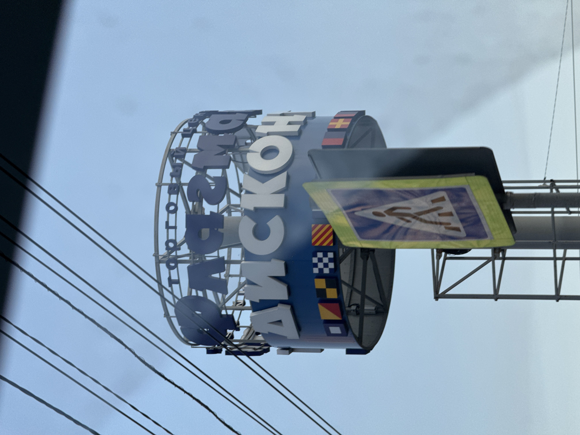 This media file captures a close-up, low-angle view of a cylindrical sign with large, bold, white and blue lettering against a pale blue sky. The sign appears to be a commercial or informational display, possibly for a business or an event. The lettering is in Cyrillic, with prominent letters like "П", "А", "М", "С", "К", "О", and "Н" visible. The sign is adorned with a series of small flag-like symbols in various colors and patterns, arranged horizontally. To the right of the main sign, a pedestrian crossing sign is visible, depicting a white pedestrian silhouette on a blue background, bordered by a yellow and green frame. The scene is dominated by the sign and the sky, with a few dark power lines running diagonally across the lower portion of the image. The overall impression is one of an outdoor urban setting. The time of day appears to be daytime, and the weather seems overcast or hazy, given the muted light and the soft appearance of the sky. There are no people visible in the frame. The orientation of the sign suggests it is a large structure, likely mounted on a pole or frame, as indicated by the metal scaffolding visible behind it. The image is somewhat blurry, particularly around the edges, suggesting it was taken with a handheld camera. The context provided places this scene in Izhevsk, Russia.