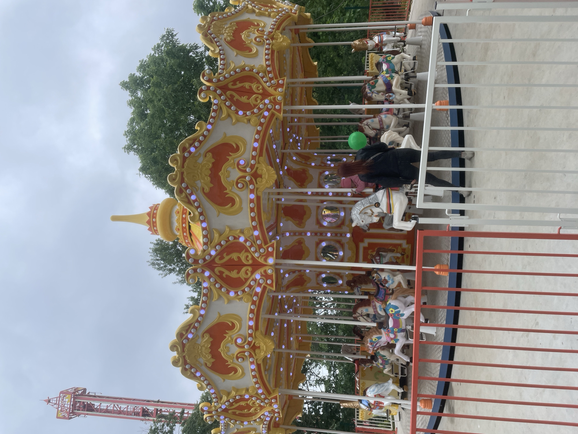 The image depicts an outdoor amusement park scene in Vladivostok, Russia, under an overcast sky. A large, ornate carousel is the central subject, featuring a multi-tiered canopy adorned with gold, orange, and white scrollwork and numerous small, glowing blue lights. Beneath the canopy, various carousel horses and at least one swan figure are mounted on poles, appearing stationary. These figures are predominantly white, brown, and multicolored.
In the foreground, a person with reddish-brown hair, viewed from behind, wears a dark jacket and dark pants, holding a bright green balloon in their left hand. This individual stands near a white metal fence that partially encloses the carousel area. To the left of this person, a figure in a pink hat is partially visible on one of the carousel horses. The carousel itself is encircled by a red metal fence.
In the background, on the far left, a tall, red and yellow amusement ride resembling a drop tower extends high into the cloudy sky. Green deciduous trees are visible behind the carousel and the distant ride, indicating a park setting. The ground surrounding the carousel is a light-colored paved surface. The carousel appears to be currently inactive.