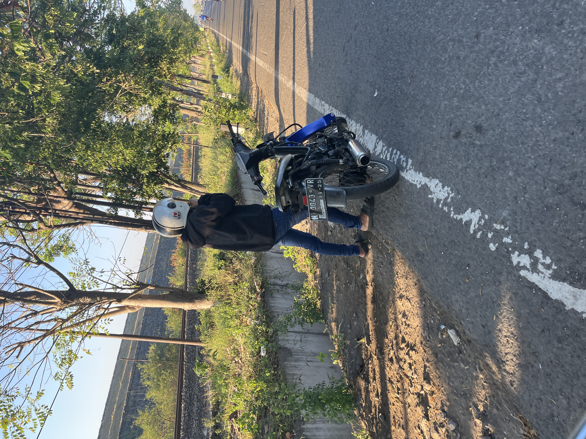 In Siring, Indonesia, a person wearing a white helmet with an "I ❤️ CR7" sticker is standing next to a dark-colored motorcycle on the side of a paved road. The motorcycle has a license plate that reads "N 3043 VR". The person is dressed in a black hooded jacket and blue jeans, with one foot on the ground and the other resting on the motorcycle's footpeg. Behind the person and the motorcycle, there is a concrete embankment with overgrown green vegetation. Beyond the embankment, railroad tracks are visible, with a slope of earth and grass in the background. A line of trees with green leaves runs along the right side of the road, and other people on bicycles can be seen further down the road in the distance. The sun is casting long shadows on the road, indicating it is either early morning or late afternoon.