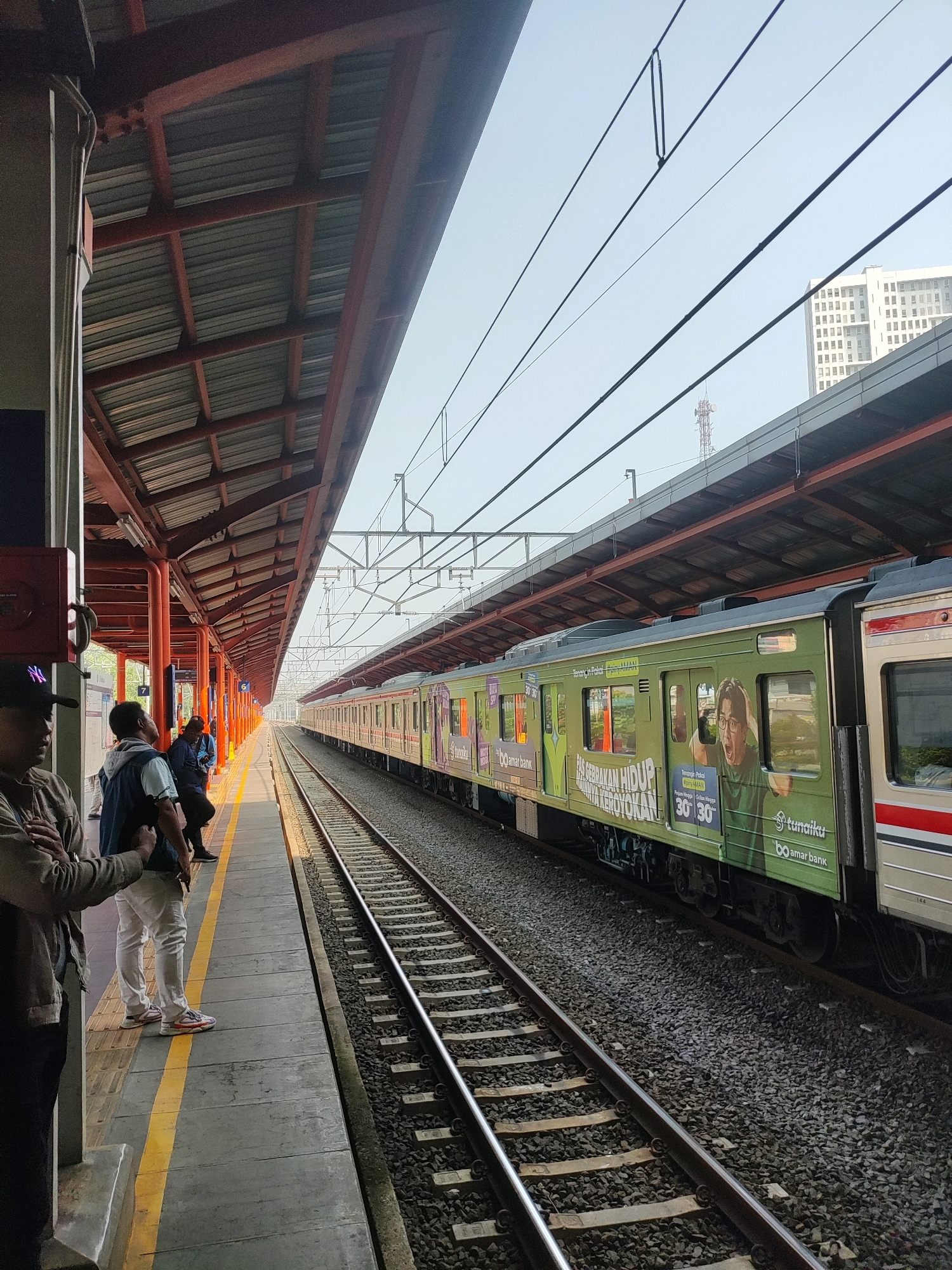 A train is stopped at a station platform in Bekasi, Indonesia. The train is light green with advertisements on its side, one of which features a man with his mouth open in surprise. The station has a covered platform with a reddish-orange support structure and a corrugated metal roof. People are standing on the platform, waiting for the train. In the background, there is a tall building and power lines. The train tracks are visible in the foreground, with ballast stone underneath. The sky is a light blue, indicating daytime.