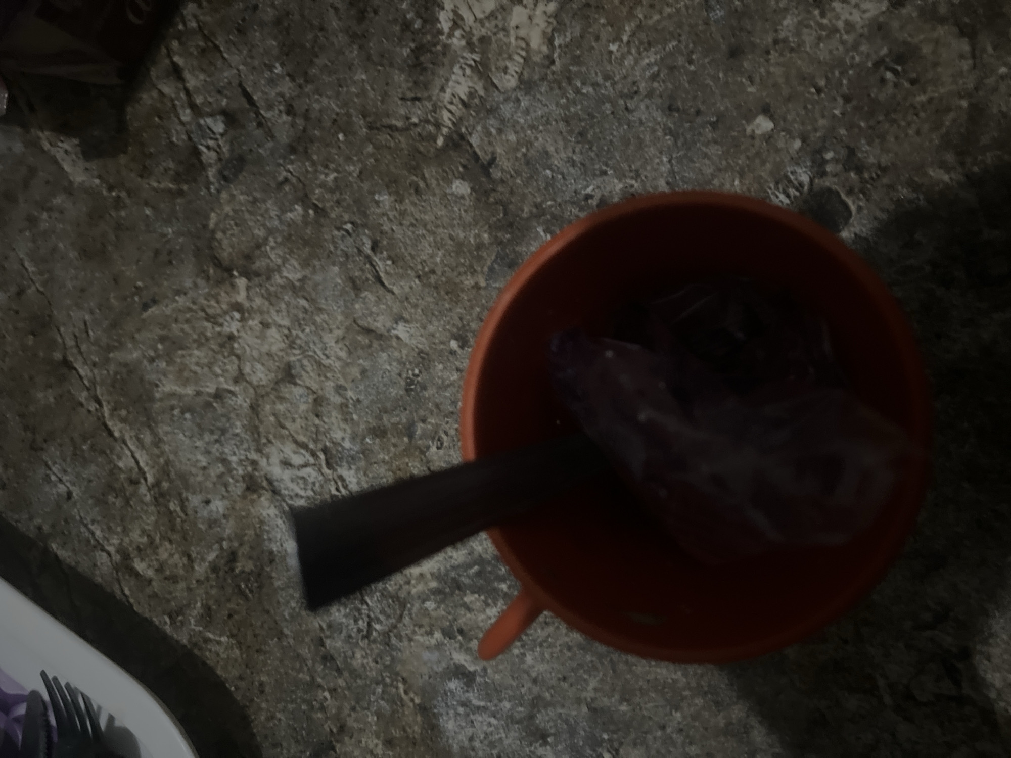 The image depicts an overhead view of an orange bucket placed on a rough, textured surface that appears to be concrete or stone. Inside the bucket, there is a dark, elongated object, possibly a stick or a handle, protruding from a transparent plastic bag. The bag contains what looks like a piece of meat or food item, with a reddish-purple hue, wrapped in plastic. The surrounding environment is dimly lit, with the ground showing cracks and variations in color. The upper left corner of the image shows a white plate with a fork and what appears to be a purple flower or food item. This scene is likely a domestic or outdoor setting in Jalingo, Nigeria, given the context of the prompt, though no specific Jalingo landmarks are visible.