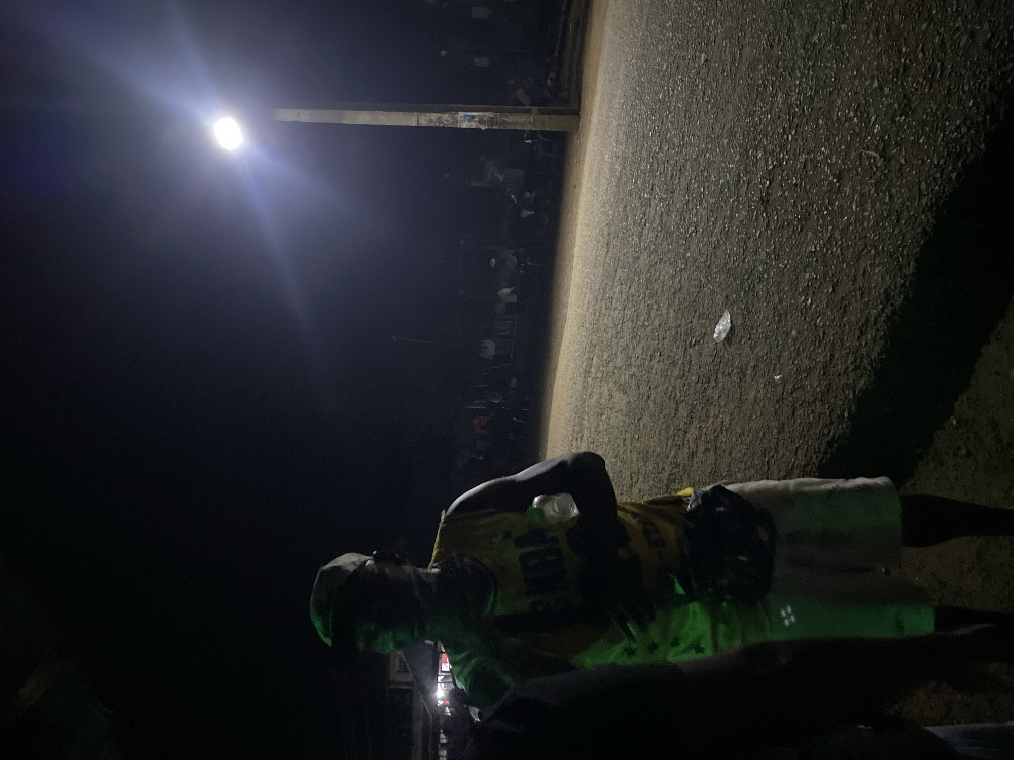 The image depicts a street scene at night in Agba Umana, Nigeria. A person wearing a yellow jersey and light-colored shorts is standing on an asphalt surface. A street light illuminates the area. In the background, there is a gathering of people. A plastic bottle is visible near the person.