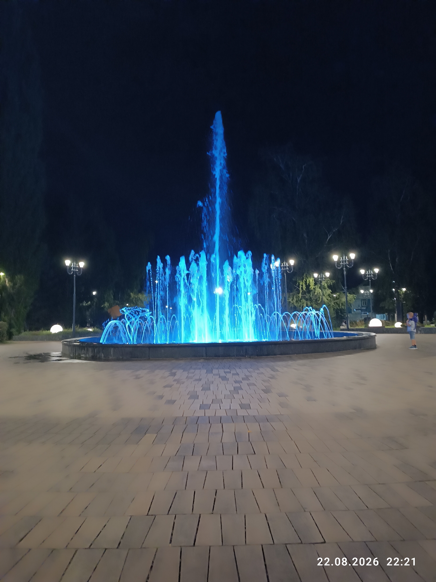 A wide-angle, eye-level, nighttime photograph shows a brightly lit fountain in the city of Sterlitamak, Russia. The fountain is a central feature, with streams of blue illuminated water shooting upwards and outwards, creating a dynamic and vibrant display. The fountain is situated in a paved plaza with a patterned brickwork surface.
Surrounding the fountain are several ornate streetlights, each with multiple bulbs, casting pools of light on the plaza. In the background, silhouetted trees create a dark backdrop against the night sky. On the right side of the image, a young boy, wearing a light blue shirt and dark shorts, stands facing the fountain. He appears to be observing the water display. Further in the background, indistinct figures and structures can be seen, including some spherical white lights along the edge of the plaza. The timestamp "22.08.2026 22:21" is visible in the bottom right corner of the image.