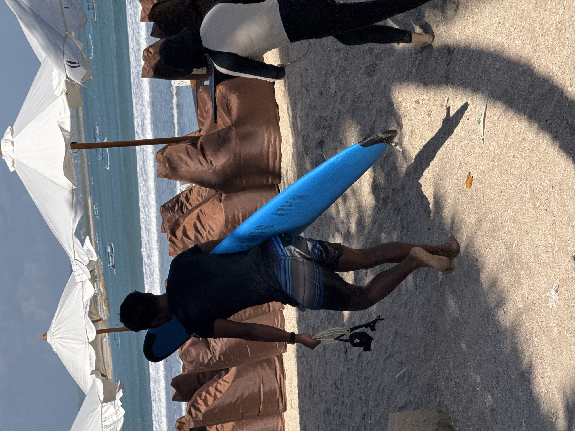 Two people are on a sandy beach in Kuta, Indonesia. In the foreground, a person wearing a black t-shirt and striped shorts walks away from the camera, carrying a bright blue surfboard. The person's left hand is holding a coiled leash with a black strap. In the middle ground, a second person wearing black leggings and a long-sleeved shirt with light-colored panels walks towards the right, also appearing to be heading towards the water. Behind the people, large brown beanbag chairs are arranged on the sand, and white beach umbrellas are set up. The ocean is visible in the background, with gentle waves and several small boats in the distance. The sky is partly cloudy.