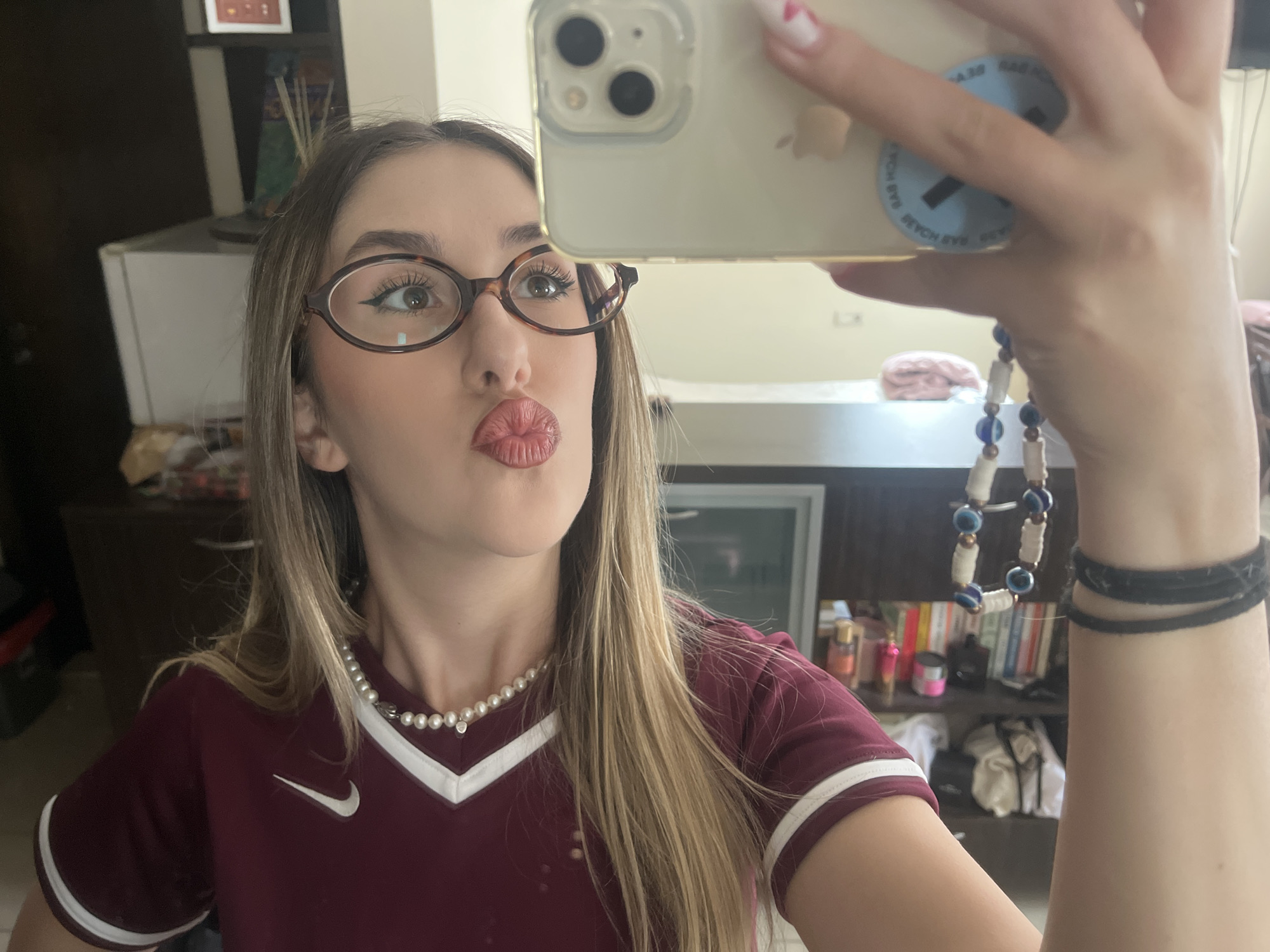 A woman takes a selfie in an interior space. She wears tortoise-shell glasses, a maroon shirt with white trim and a Nike swoosh, and a pearl necklace. She holds an iPhone with a phone grip on the back. She has bracelets on her wrist. Behind her is a cabinet with items on shelves. The city and country are not discernible from the image provided.