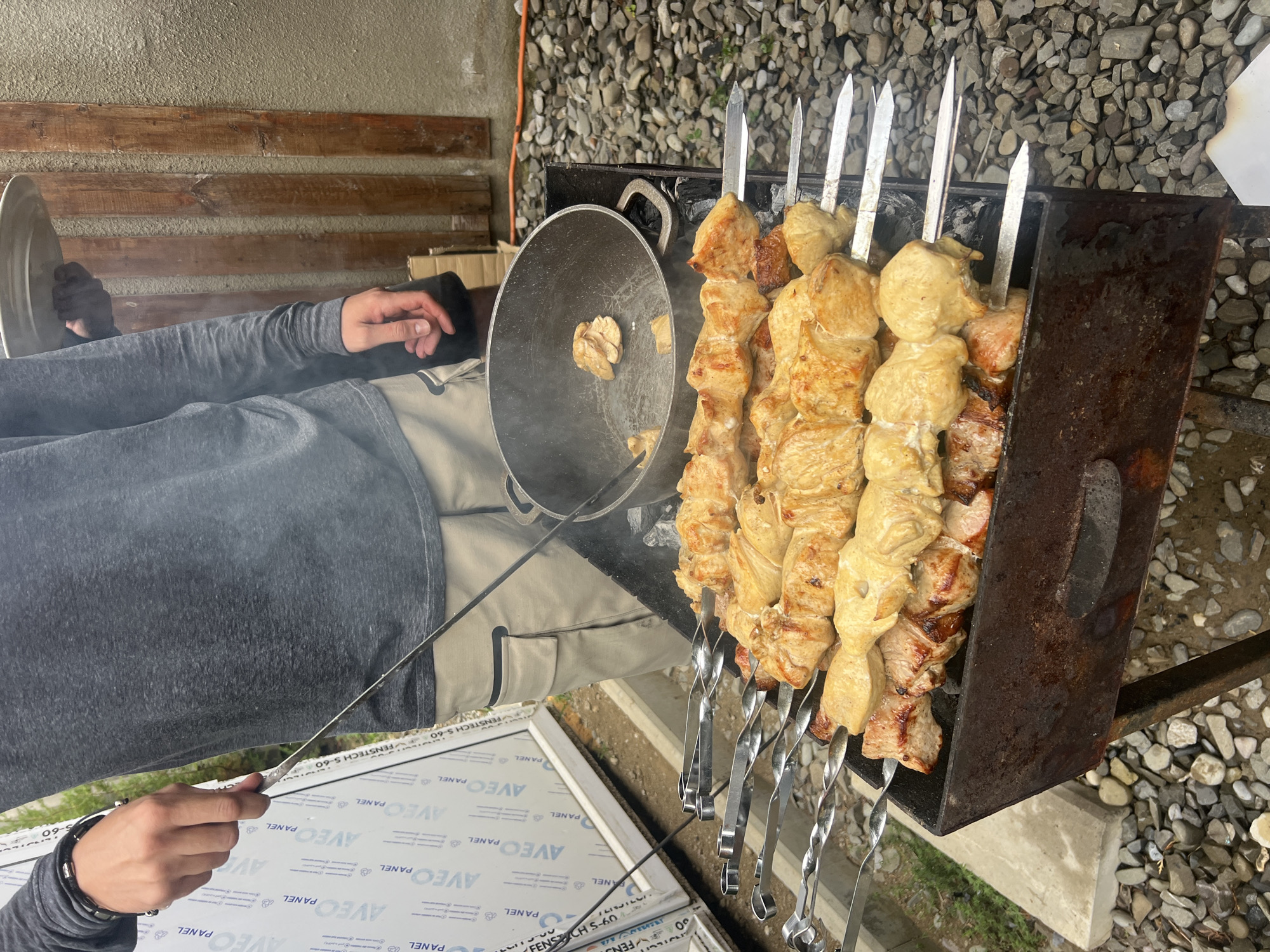 In Tsodoreti, Georgia, a person wearing a grey long-sleeved shirt and beige cargo pants is cooking shashlik (kebabs) on a charcoal grill. The shashlik, skewered with metal rods, consists of meat that appears to be chicken and pork, some of which is visibly browned from grilling. A cast-iron pot with some cooked pieces of meat sits on the grill next to the shashlik. The surrounding environment is outdoors, with a gravel ground, a concrete slab, and a wall constructed of wooden planks. In the background, there are panels with the "AVEC PANEL" logo. The scene suggests a casual outdoor cooking event.