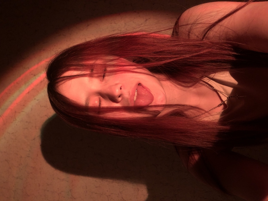 A single young female is depicted indoors, likely in the city of Фоки, Russia, though the specific address is not discernible from the image. She has long, straight, dark hair, which is heavily illuminated by a red light source, giving it a reddish hue. Her eyes are closed, and her tongue is extended outward. She is wearing a light-colored top and a silver-colored necklace with a cross pendant visible at her chest. The background is a textured, light-colored wall, also bathed in red light, with a distinct shadow of the subject's head and upper body cast to the left. Two parallel, curved streaks of red light are visible on the upper right portion of the wall. The lighting suggests an artificial red light source, potentially a projector.