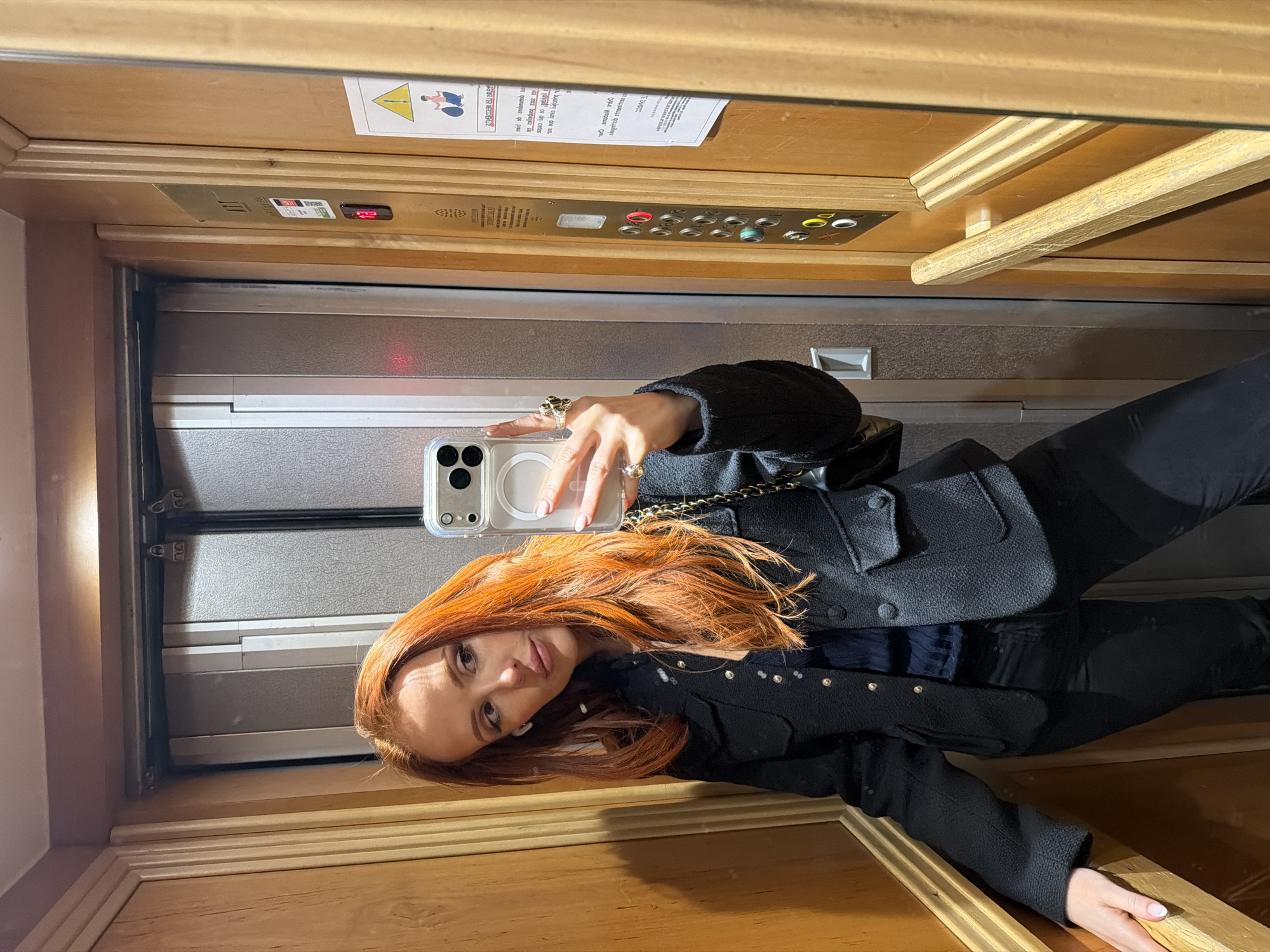 A woman with long red hair, wearing a black jacket and dark pants, is taking a selfie in an elevator. She is holding a white smartphone with a clear case. The elevator has wood paneling on the floor and ceiling, and mirrored walls. Elevator buttons and a sign with a pictogram and text are visible above her. The OCR suggests the location is Paris, France due to the French text in the sign.