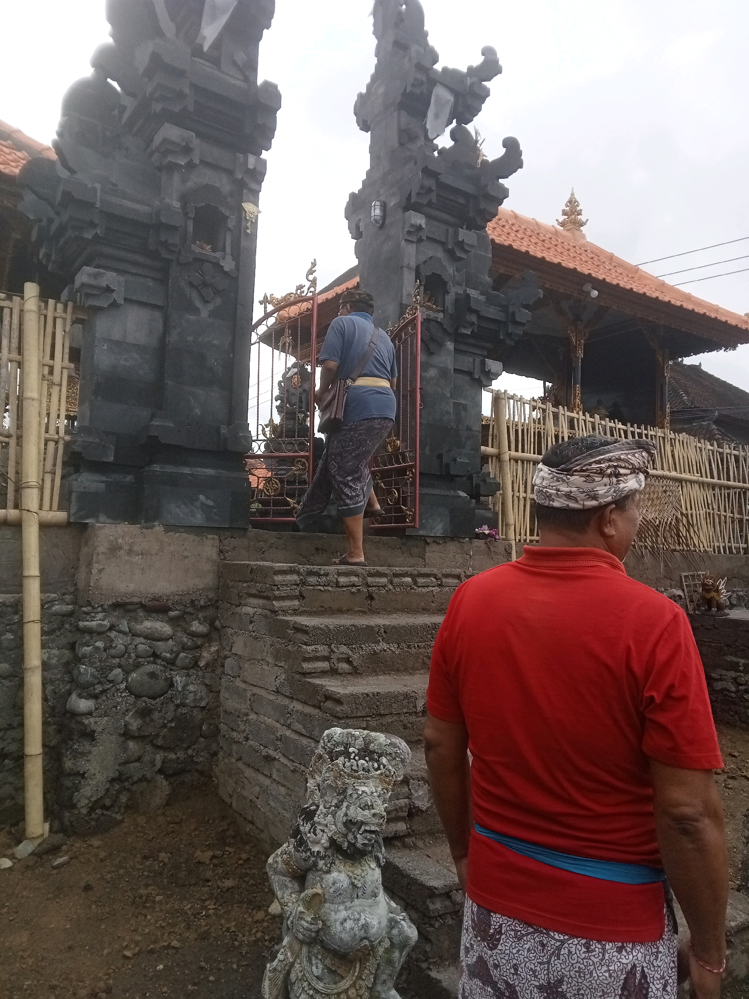 In the city of Mendoyo, Indonesia, two men stand near a temple entrance. The man in the foreground faces away from the camera, wearing a red t-shirt and a patterned sarong. A blue sash is visible around his waist. He has a patterned cloth wrapped around his head. The second man, facing left, is seen from the side and back as he walks up stone steps towards the temple. He wears a blue shirt and a patterned sarong, with a brown bag slung over his shoulder. The temple features imposing dark stone gateways and a tiled roof. Bamboo fencing is visible behind the gateway. A weathered stone statue of a mythical creature stands at the base of the steps. The ground is a mix of dirt and small stones. The sky is overcast.